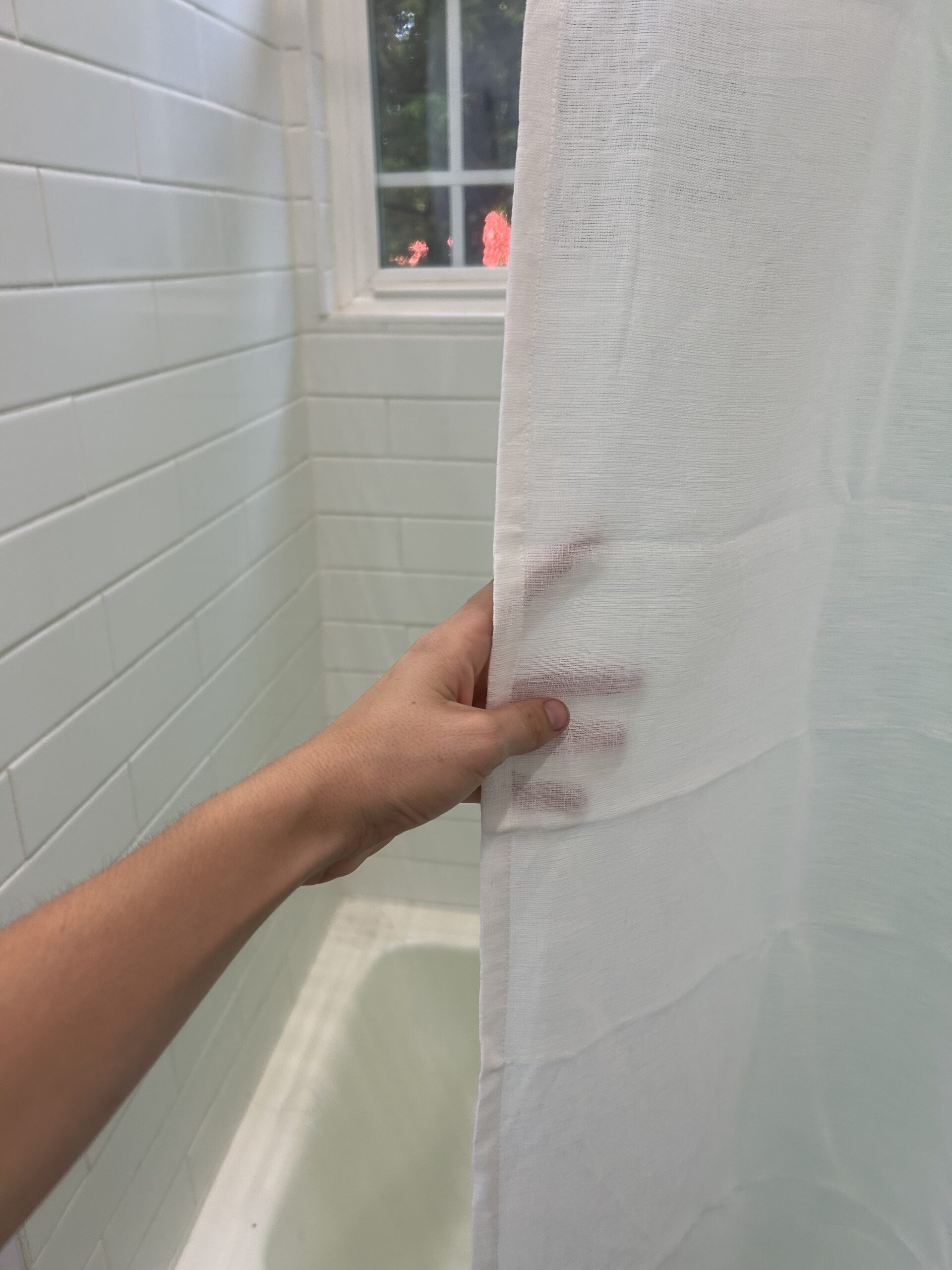 A hand holds a white shower curtain partially open in a tiled bathroom with a window above the bathtub.