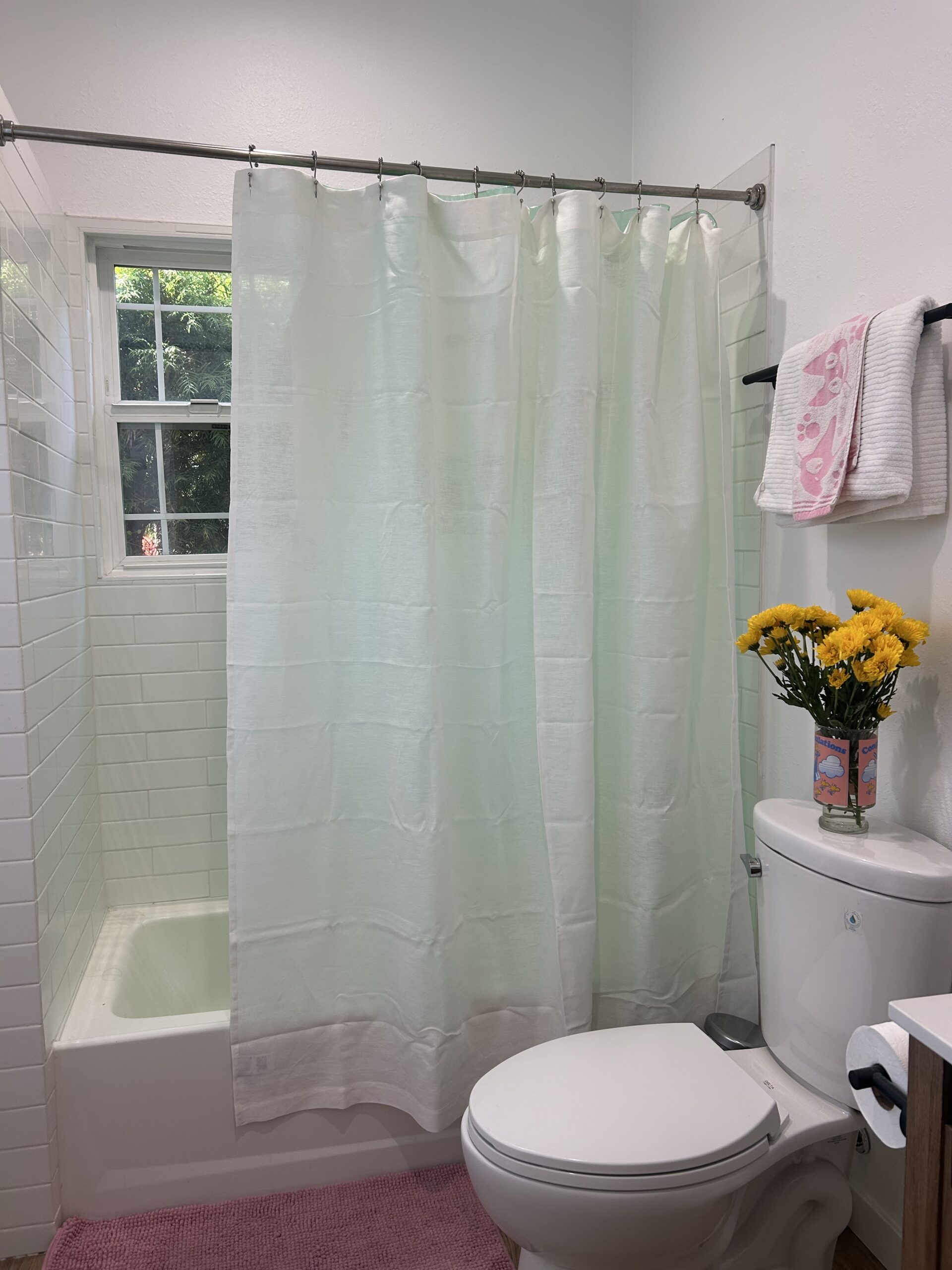 Small bathroom with a shower and white curtain, toilet, pink rug, yellow flowers in a vase, and white towels near a window.