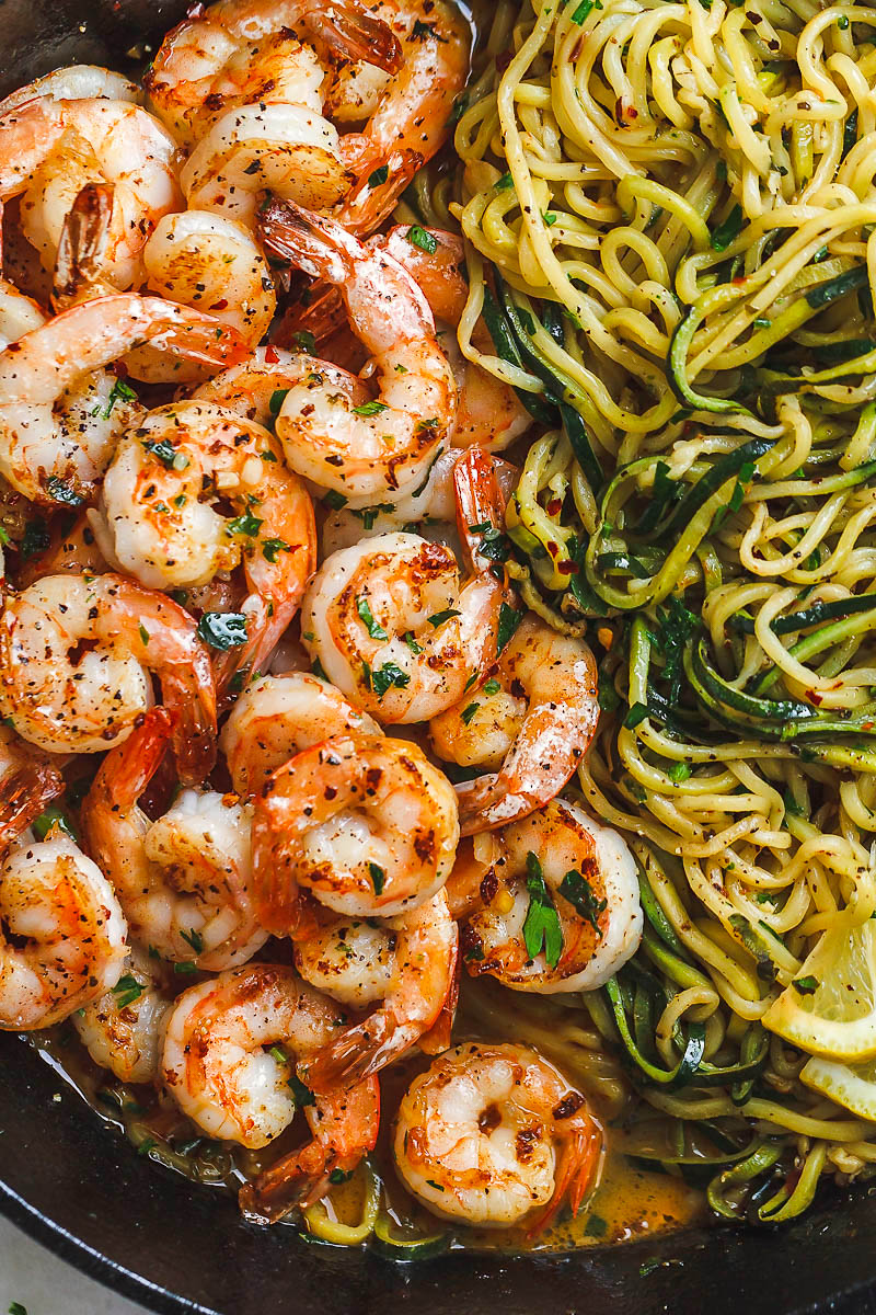 Cooked shrimp seasoned with herbs and pepper sit next to a serving of zucchini noodles in a skillet, garnished with lemon slices and parsley.