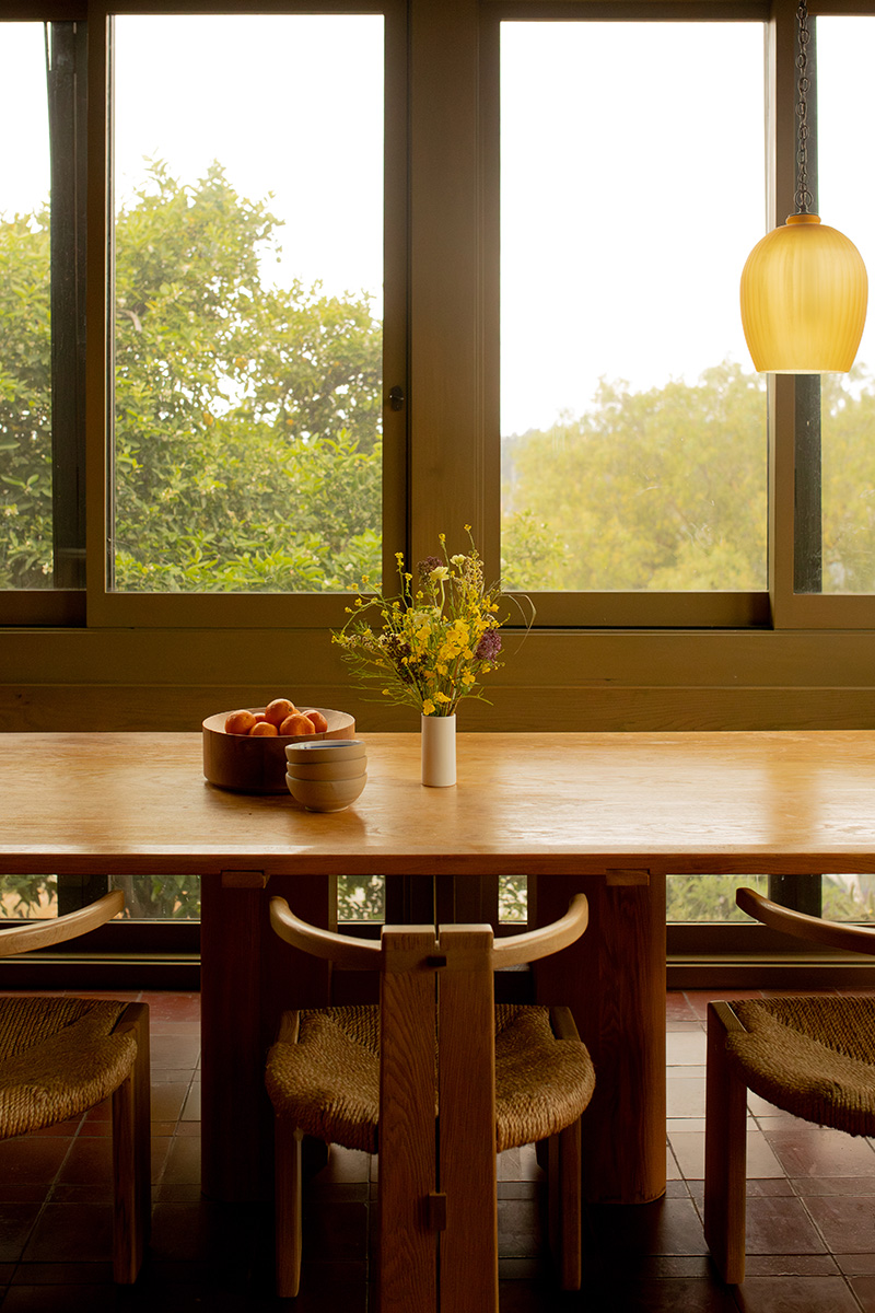 A wooden dining table with a vase of flowers and bowls of fruit, surrounded by woven chairs, sits by large windows overlooking greenery.