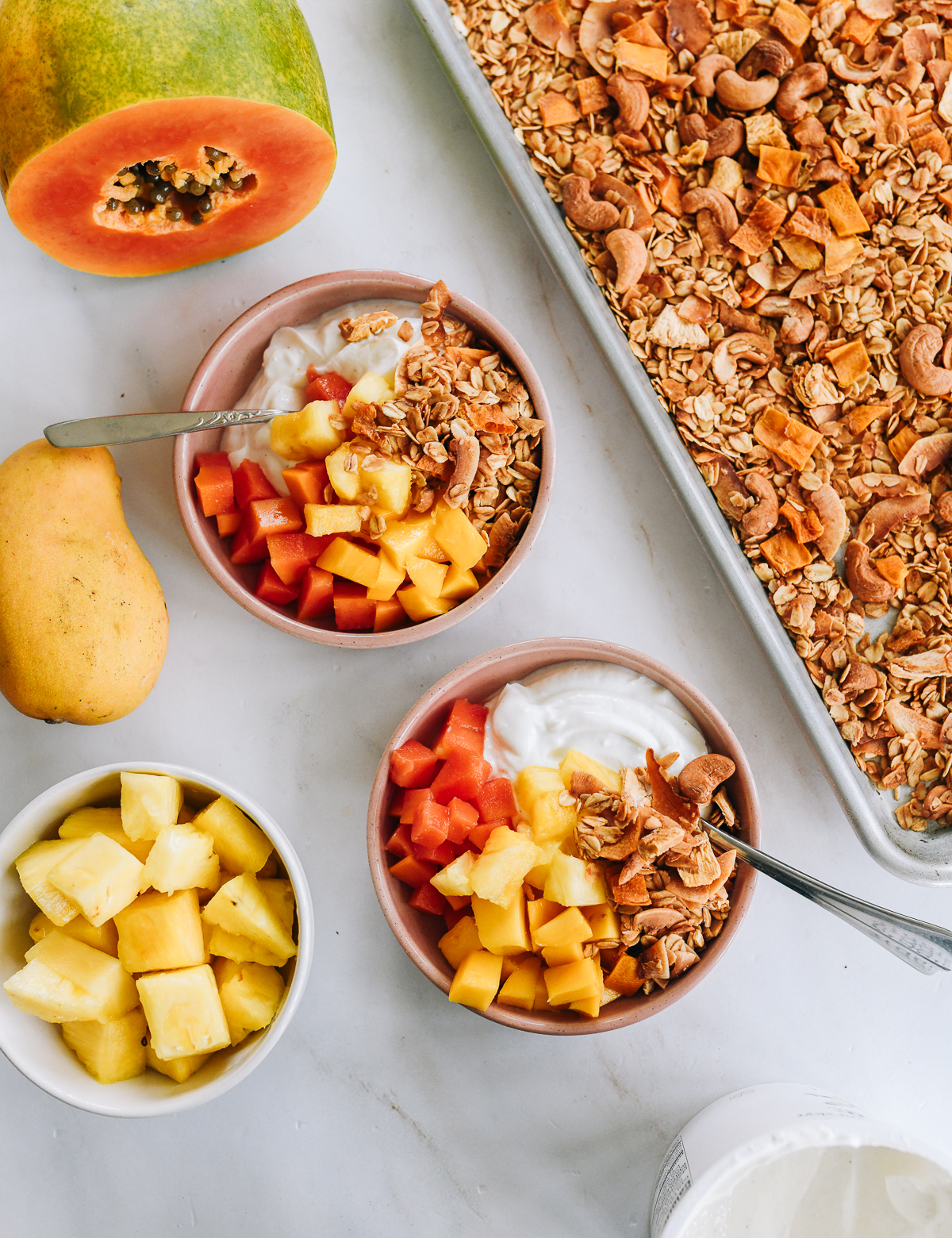 Two bowls of yogurt topped with diced papaya, mango, pineapple, granola, and coconut sit near fresh fruit, a tray of granola, and a container of yogurt on a white surface.