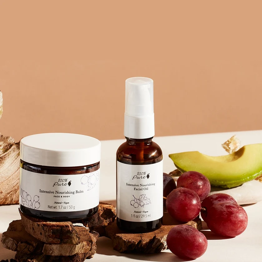 A jar of 100% Pure Intensive Nourishing Balm and a pump bottle of Intensive Nourishing Facial Oil, displayed with avocado and red grapes.