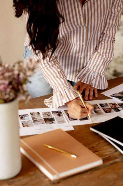 A person in a striped, long-sleeved shirt writes on a contact sheet of photographs on a wooden table.