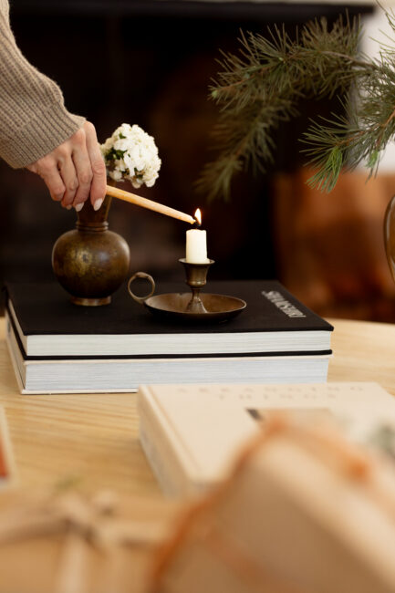 A hand uses a taper candle to light a short candle in a brass holder resting on a stack of books.