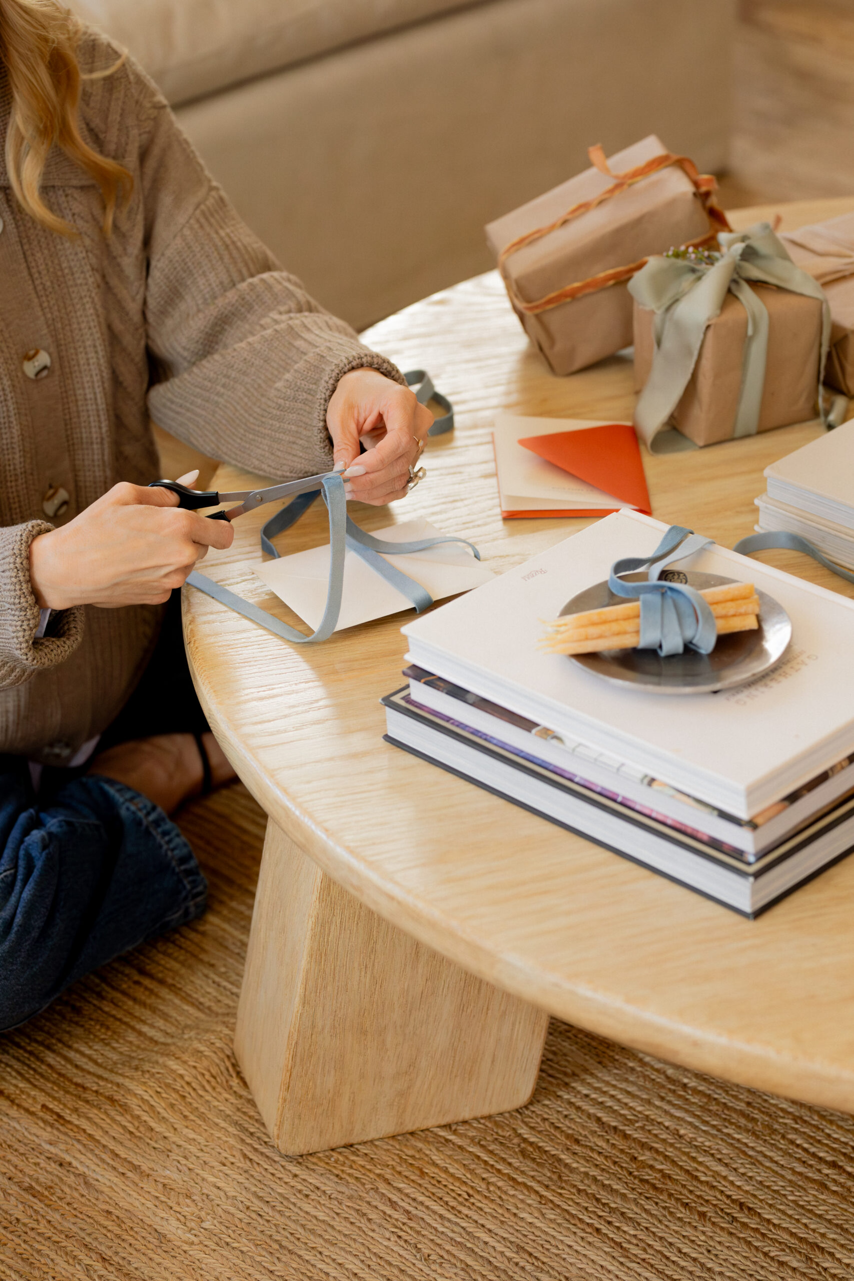 Person sitting at a round wooden coffee table, using scissors to trim a blue ribbon for a gift.