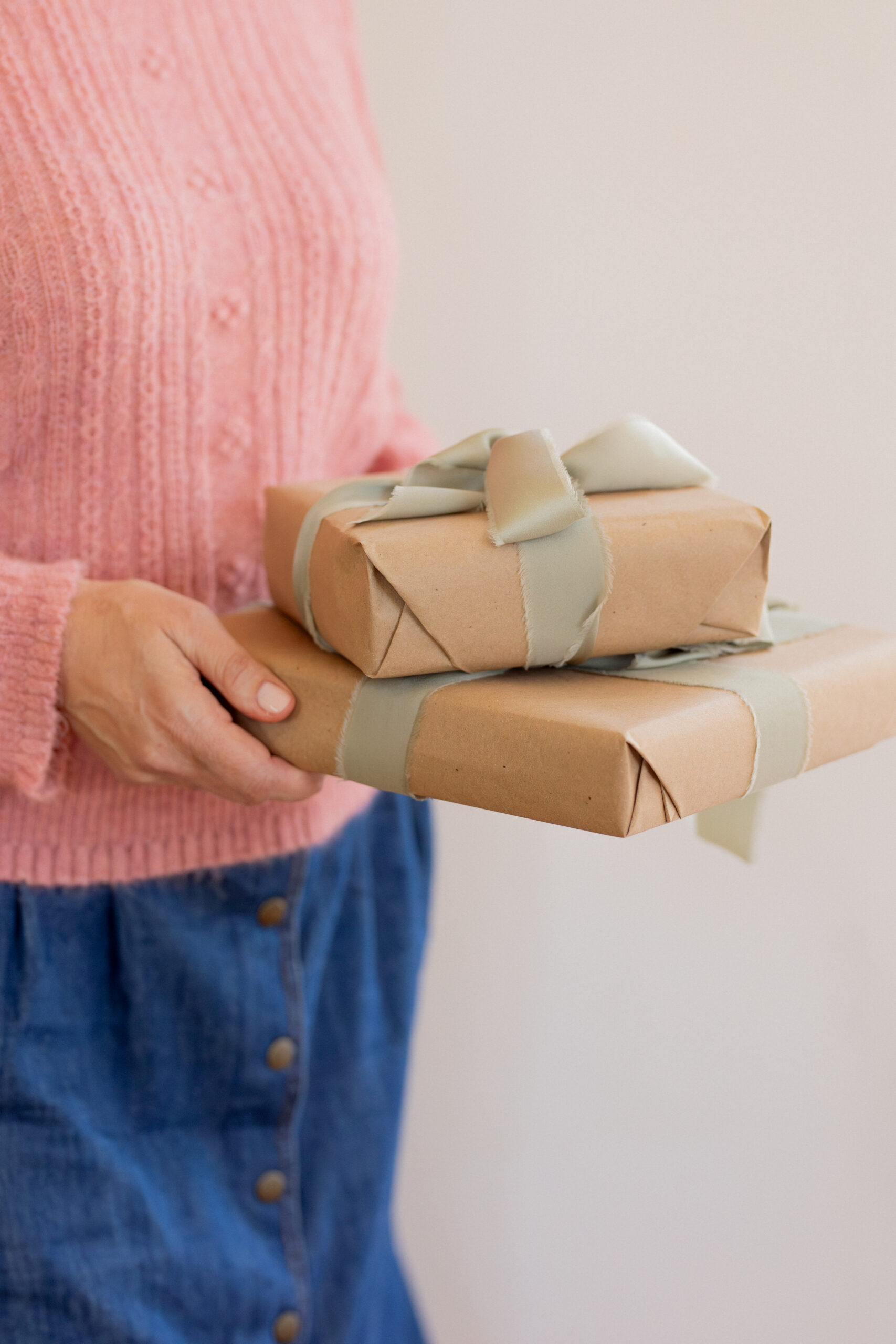A person wearing a pink sweater and blue skirt holds two brown paper-wrapped gifts tied with sage green ribbons.