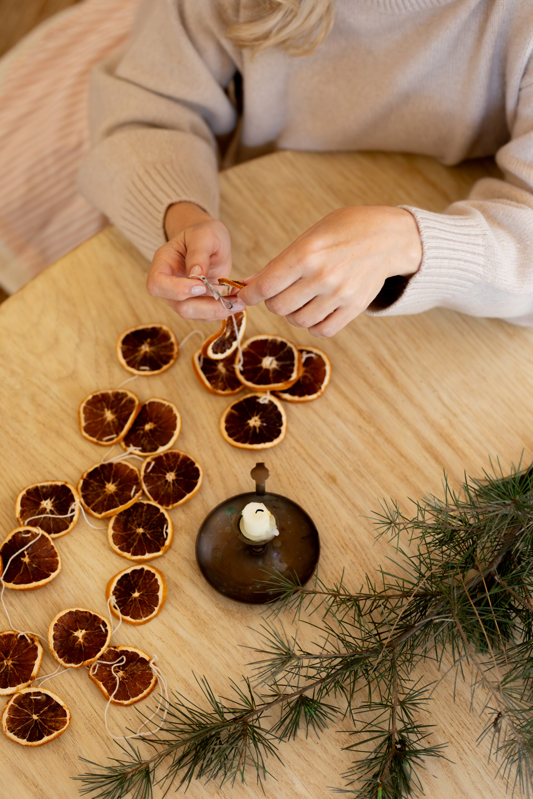 Hands threading a dried orange slice onto a string, surrounded by more slices and a small candle holder on a wooden table.