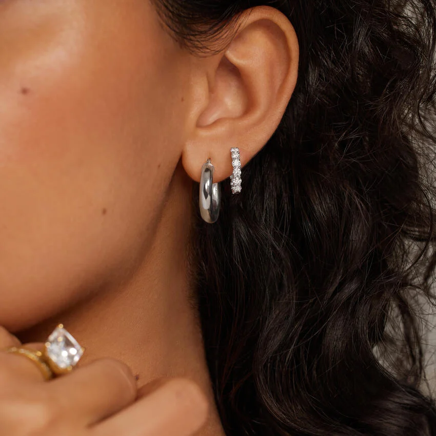 A person wearing a plain silver hoop earring and a small, diamond-encrusted hoop earring in their earlobe, with a large diamond ring visible on their hand.