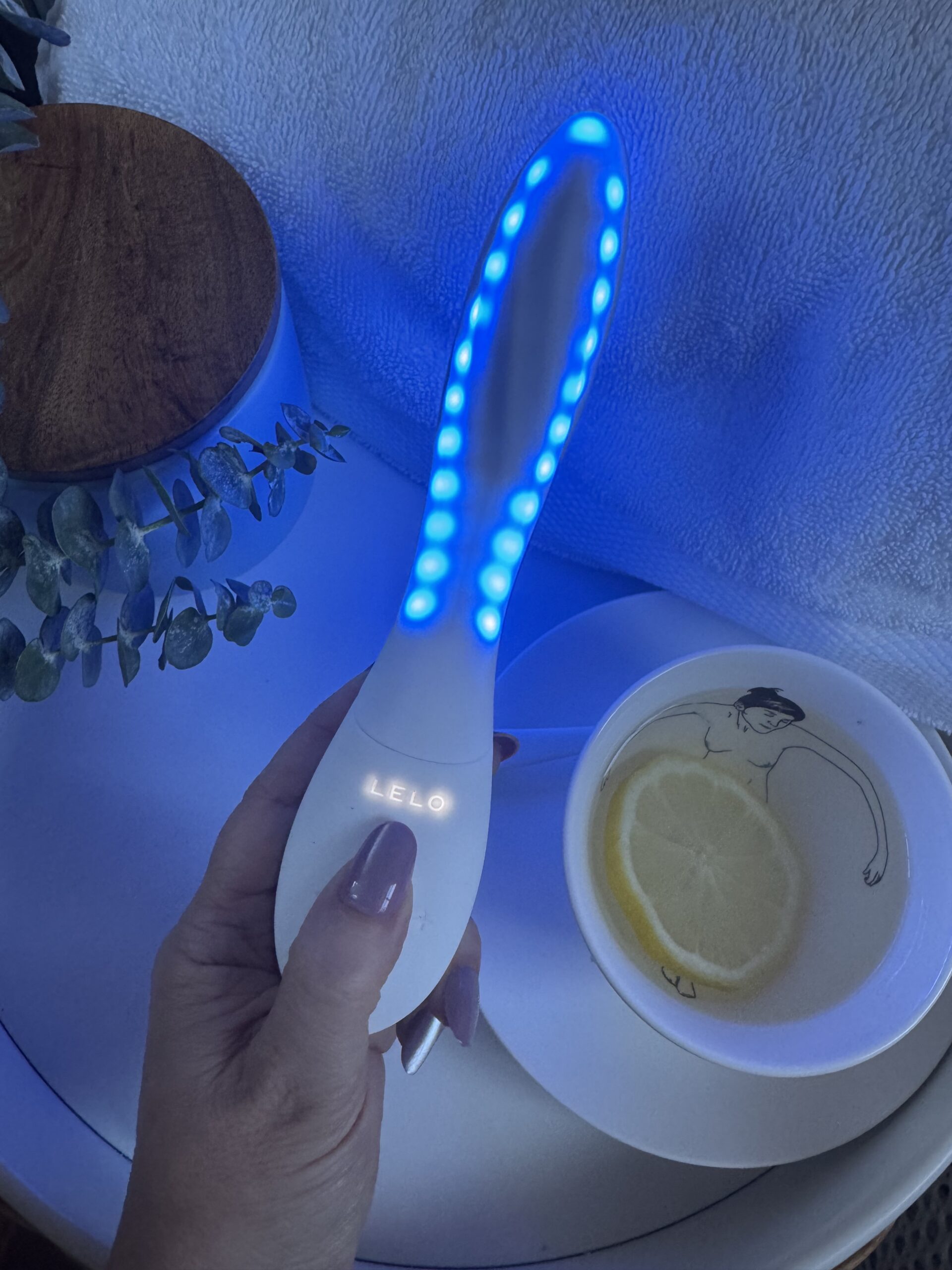 Hand holding a LELO brand LED light therapy wand next to a teacup with a lemon slice.