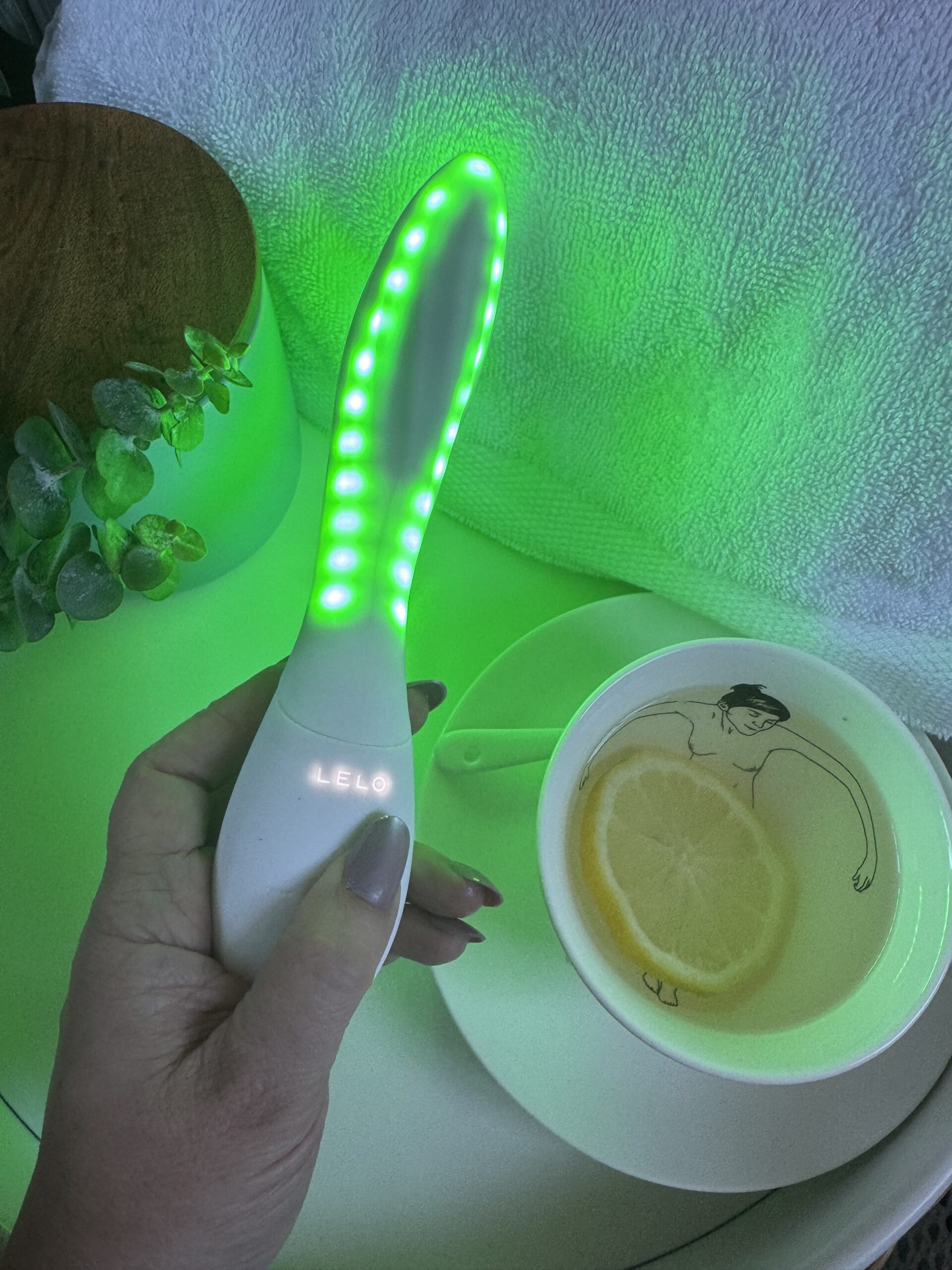 A hand holding a glowing green LELO skincare device next to a teacup with a lemon slice.