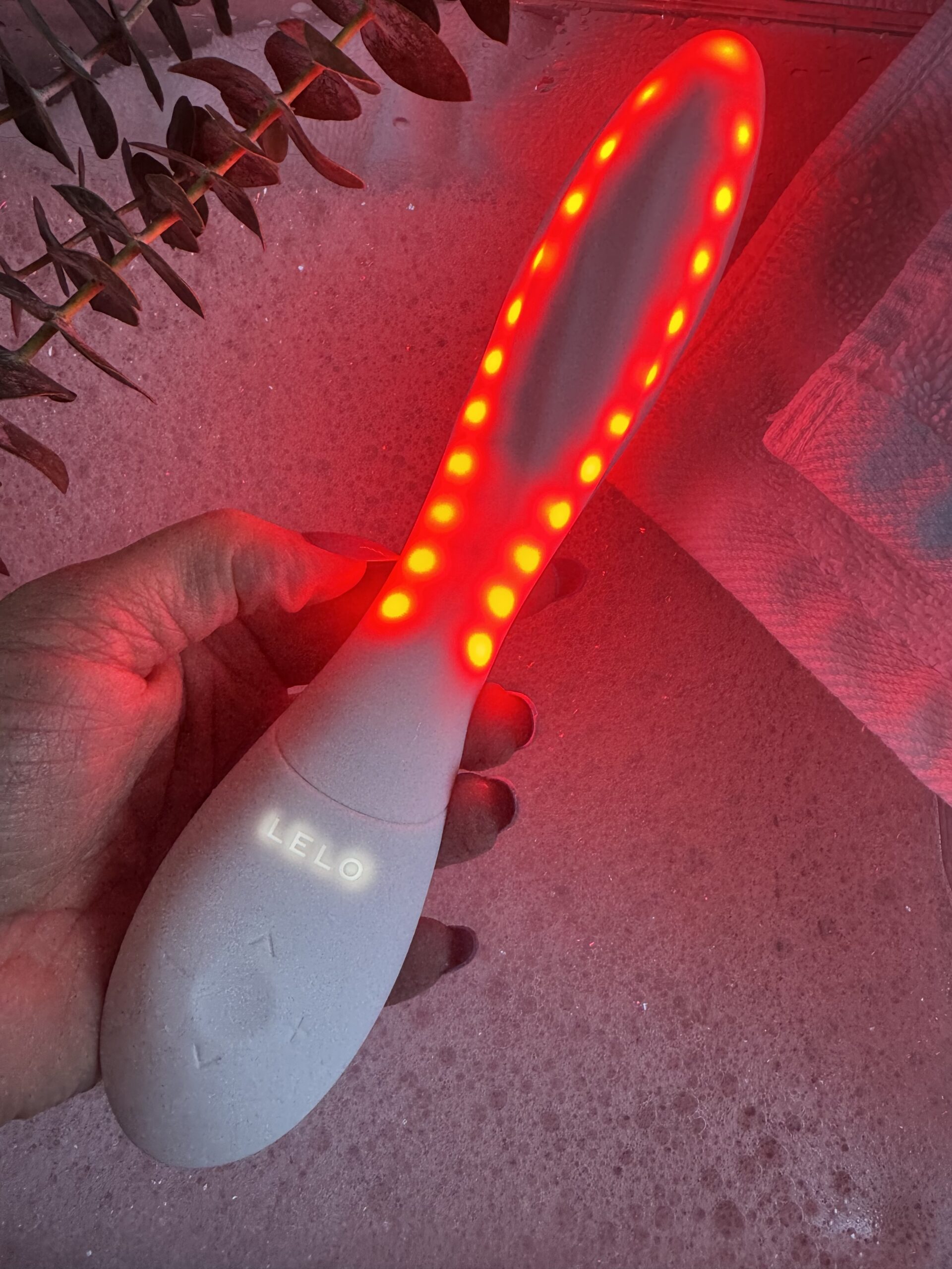 A hand holds a Lelo brand light therapy wand with red LED lights illuminated along its oval head.
