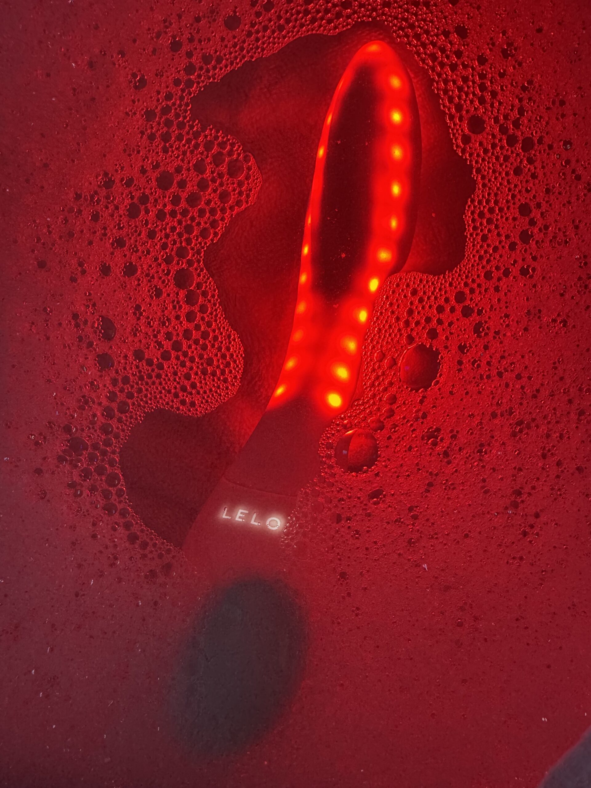 A LELO branded massager illuminated in red light, partially submerged in soapy water.
