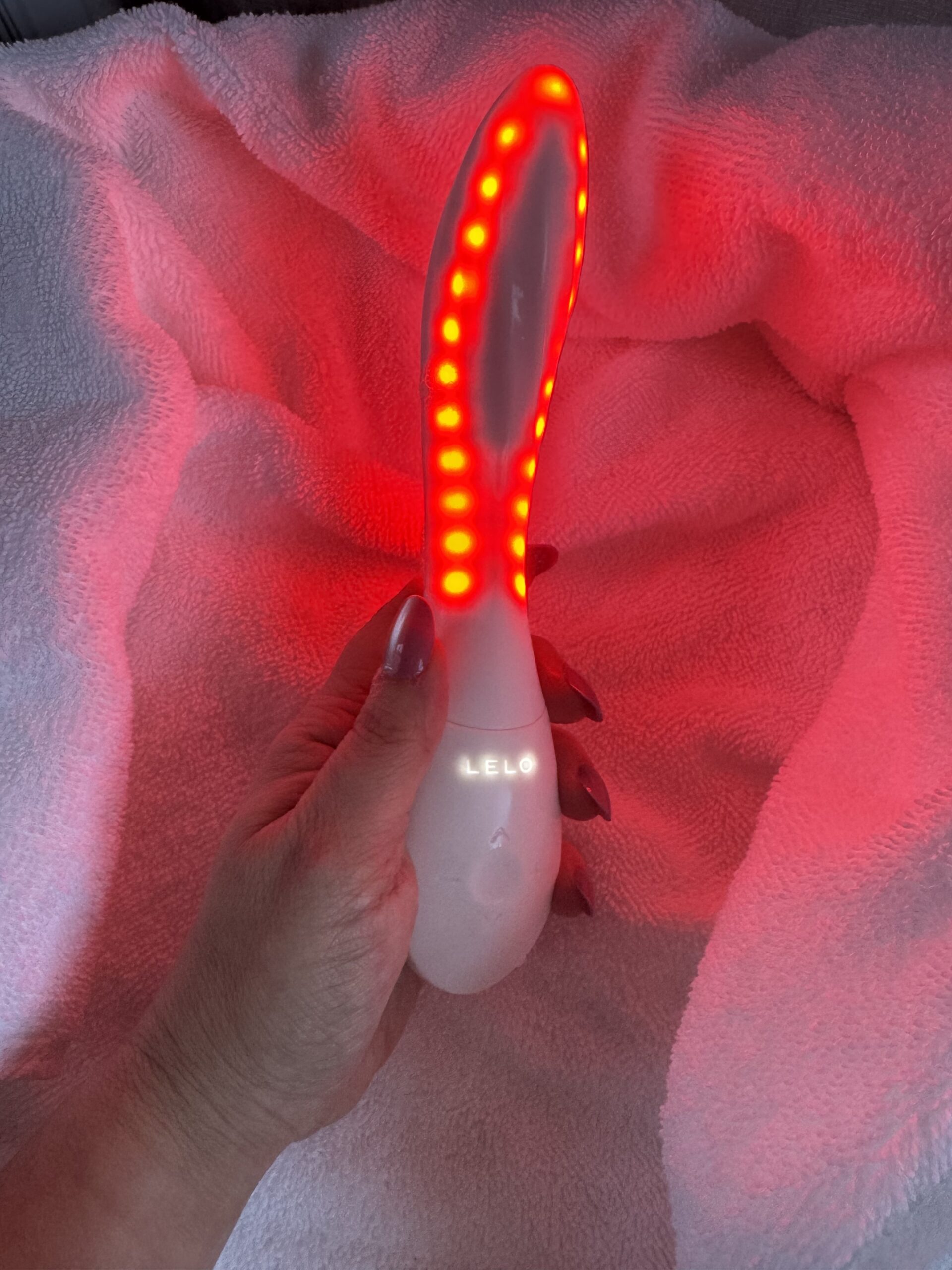 Hand holding a white, curved LELO personal massager emitting a bright red light from a row of LED indicators.