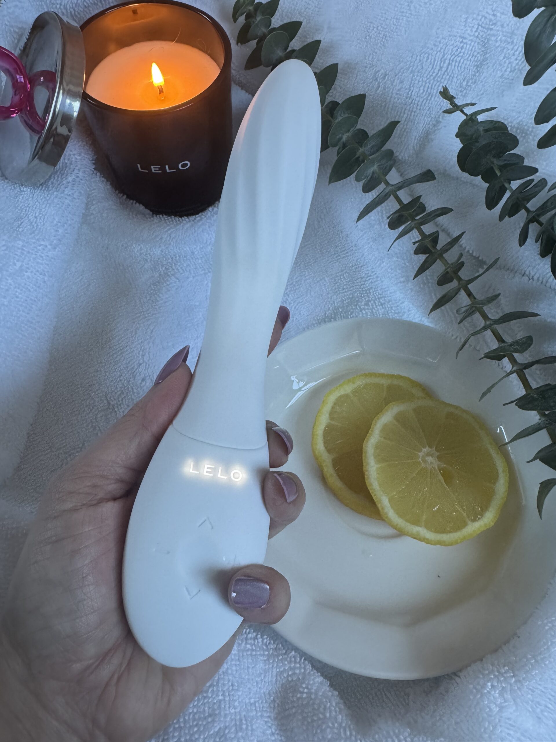 A hand holding a white LELO wellness device, with a lit candle, lemon slices, and eucalyptus sprigs on a white towel.