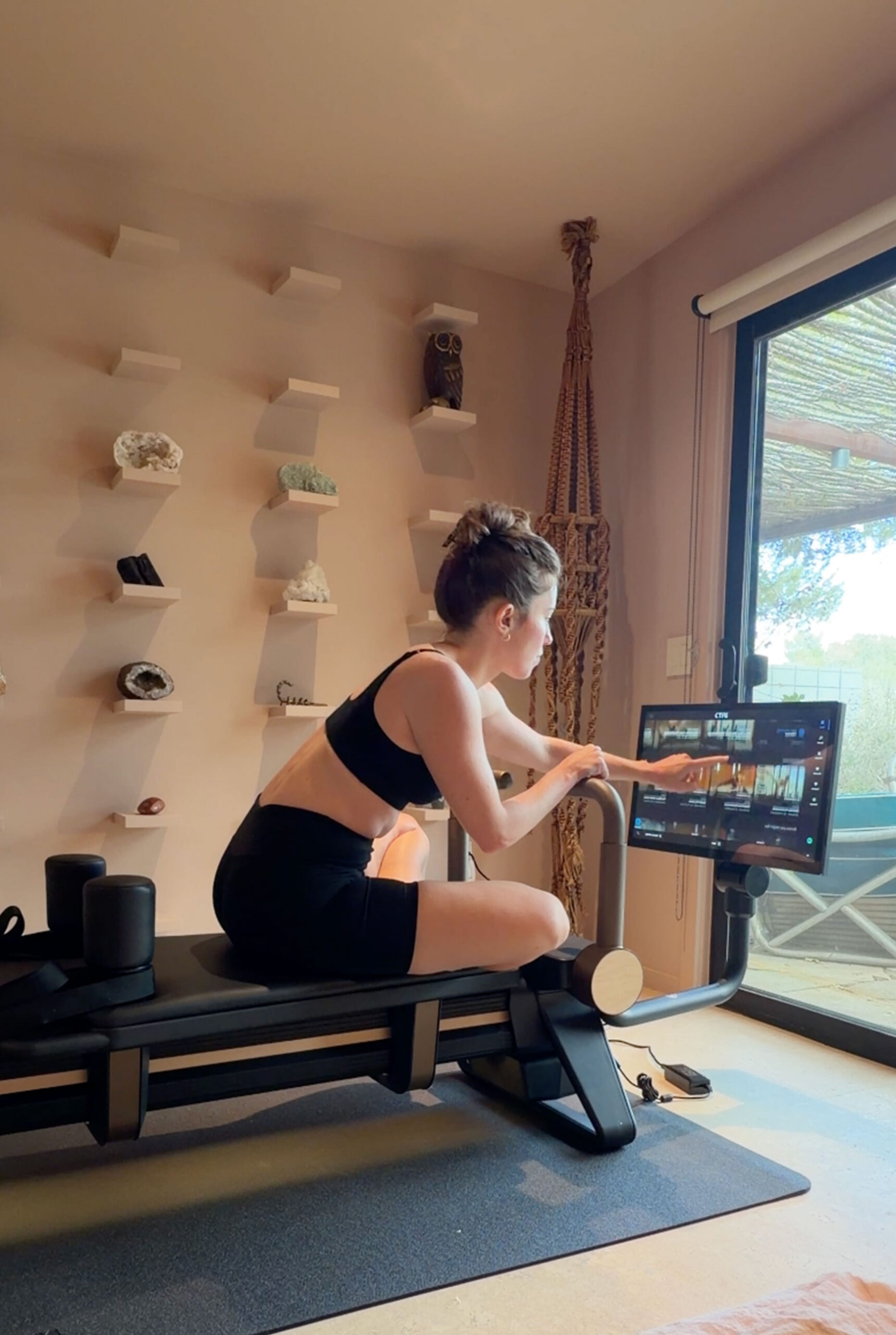 A person in athletic wear sits on a Pilates reformer, tapping the screen of a tablet attached to the machine.