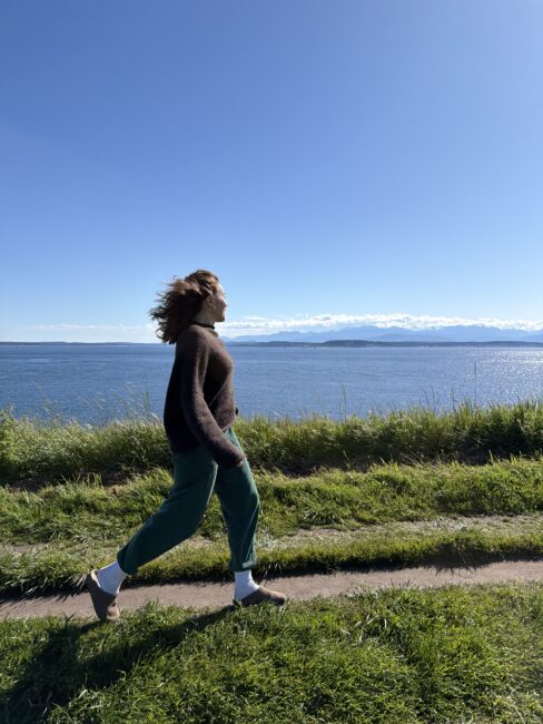 A person walking along a grassy path overlooking a bay with mountains in the distance.