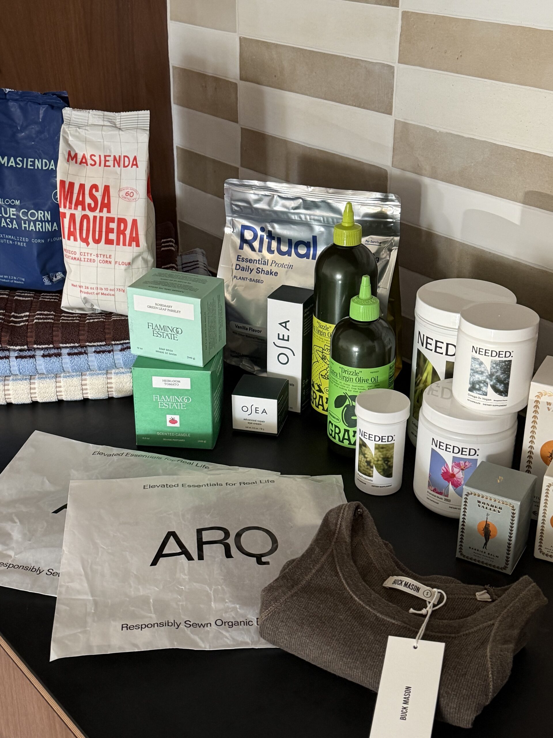 A collection of pantry items, skincare, and apparel on a counter, including Masienda masa harina, Ritual protein powder, Graza olive oil, Needed supplements, Flamingo Estate candles, and ARQ packaging.