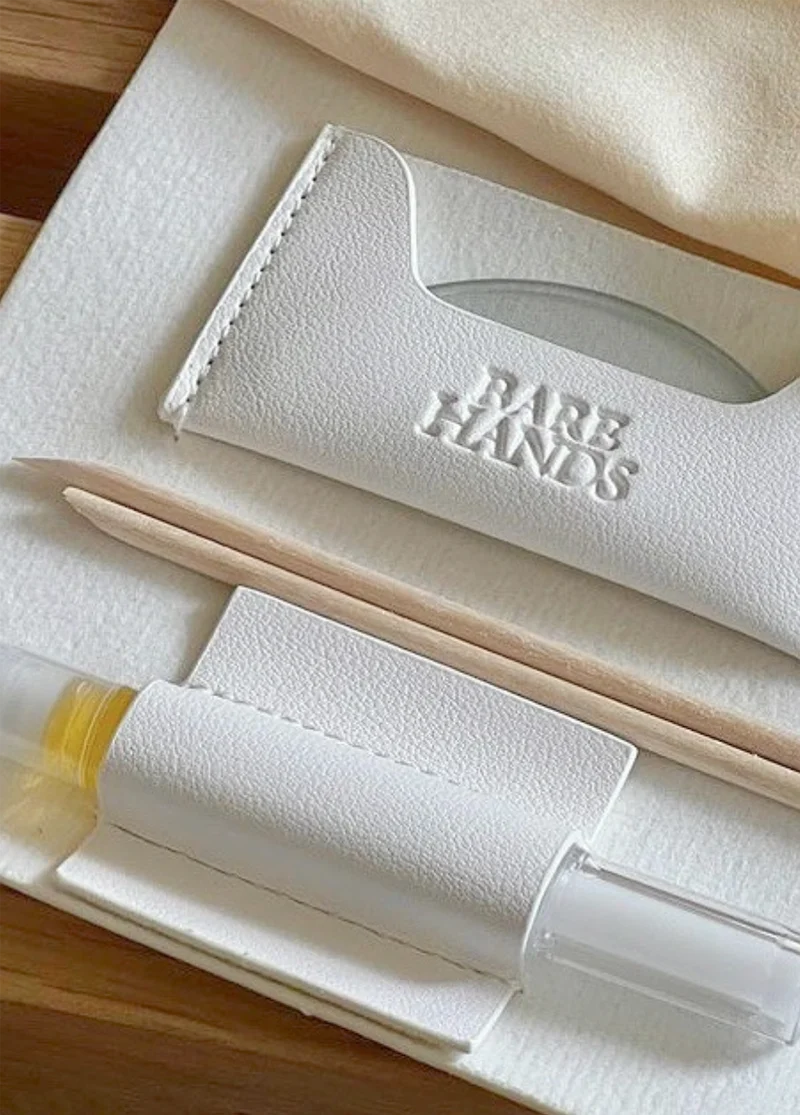 A white case embossed with “BARE HANDS” containing two wooden cuticle sticks and a small vial of nail oil.