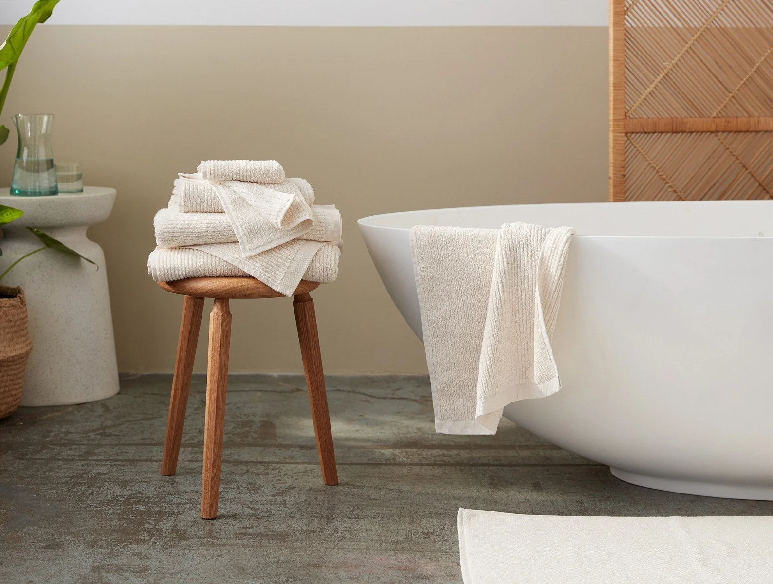 A stack of folded white towels sits on a wooden stool beside a modern white bathtub, with another towel draped over the tub.