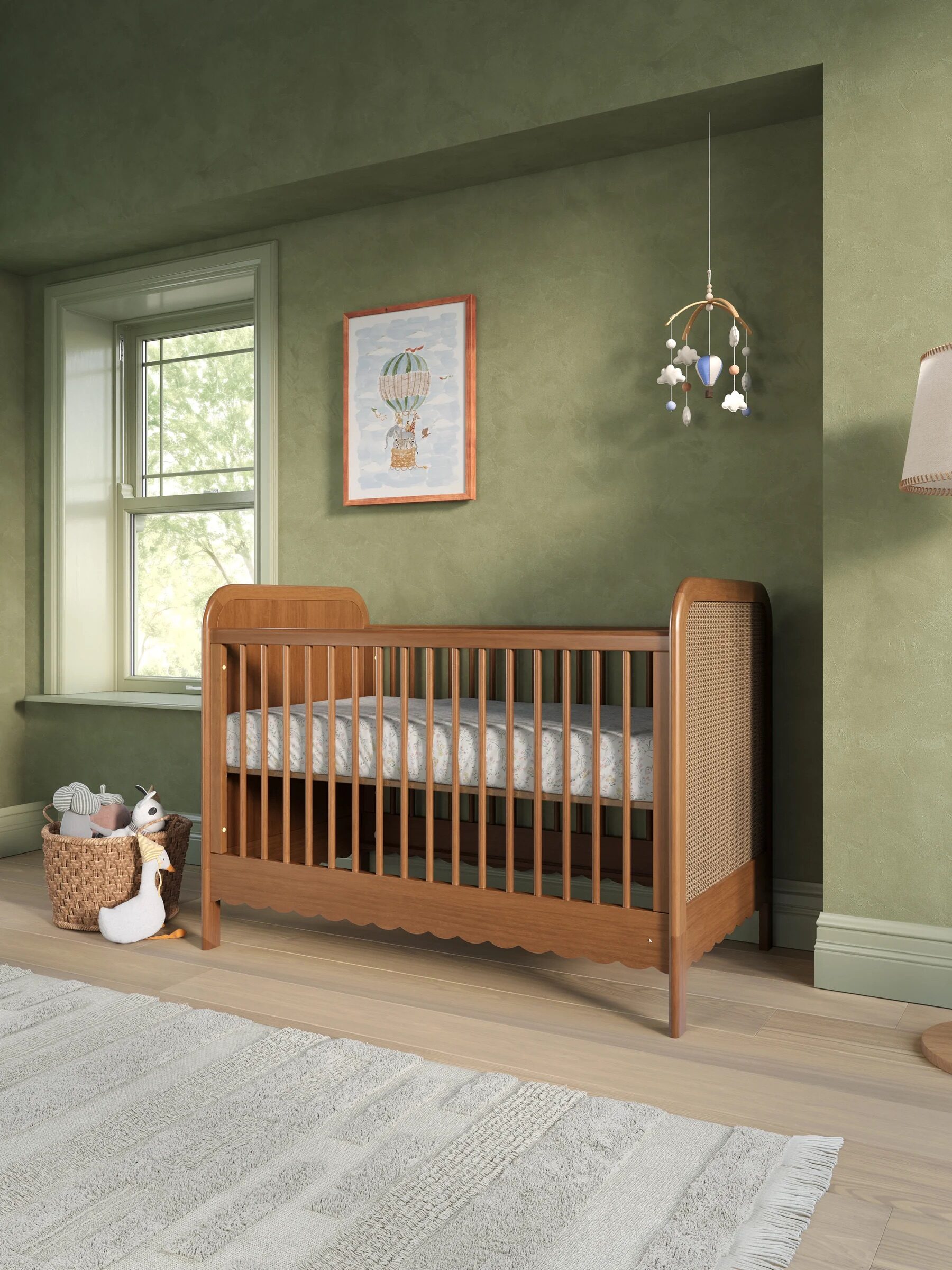 Wooden crib with scalloped trim in a nursery with green walls, a wicker toy basket, and a hot air balloon wall print.