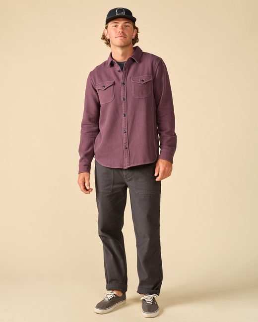 A man wearing a black baseball cap, a plum button-down shirt, dark gray pants, and gray sneakers.