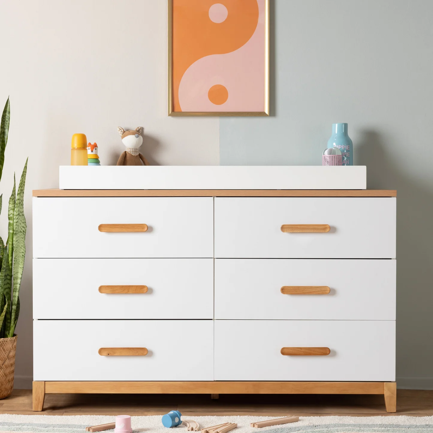 A white six-drawer dresser with wooden handles and a changing pad on top, featuring a framed abstract print on the wall above.