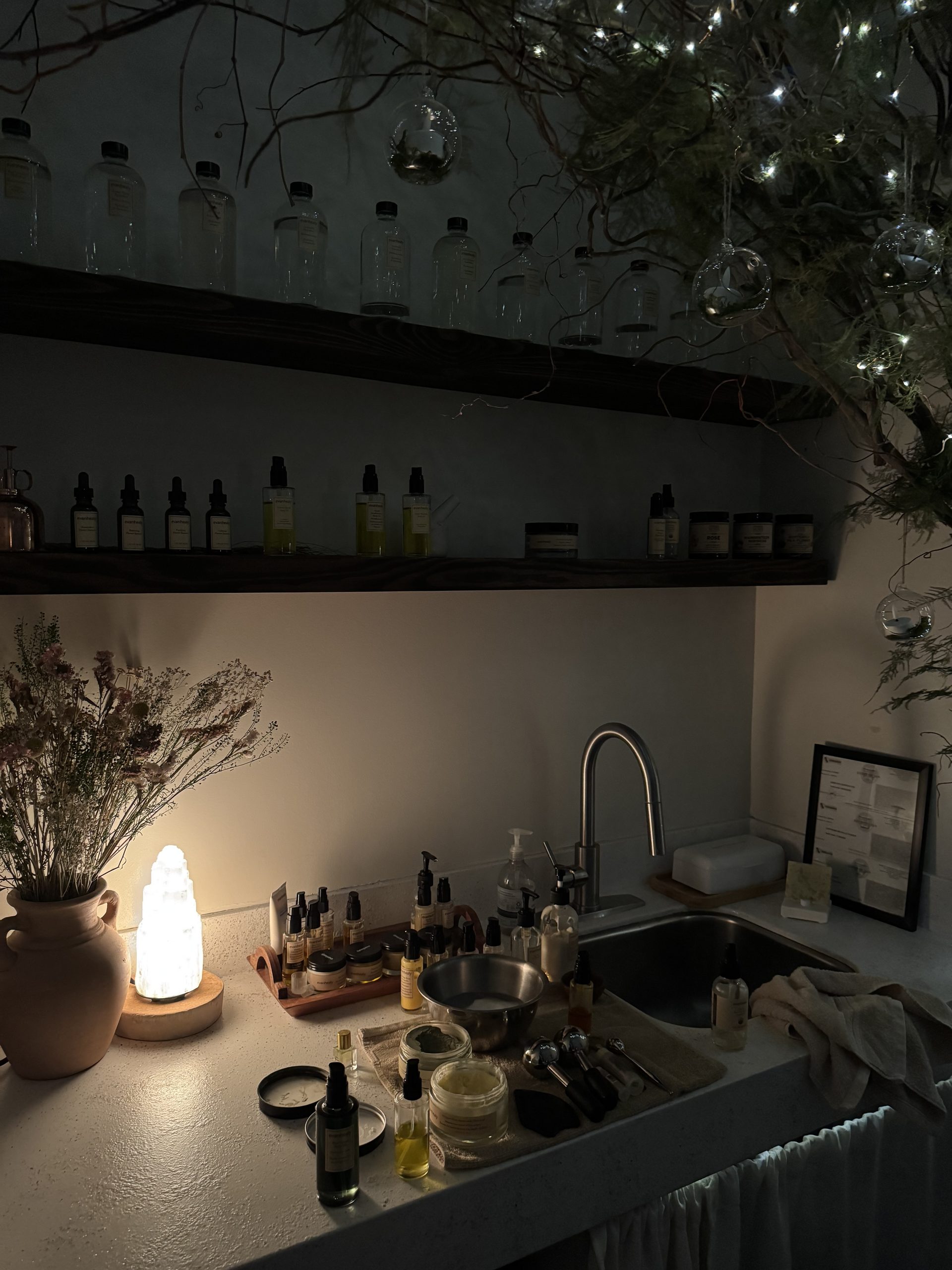 A sink area with skincare products, jars, and bottles arranged on the counter, illuminated by a glowing salt lamp and decorative fairy lights in tree branches overhead.