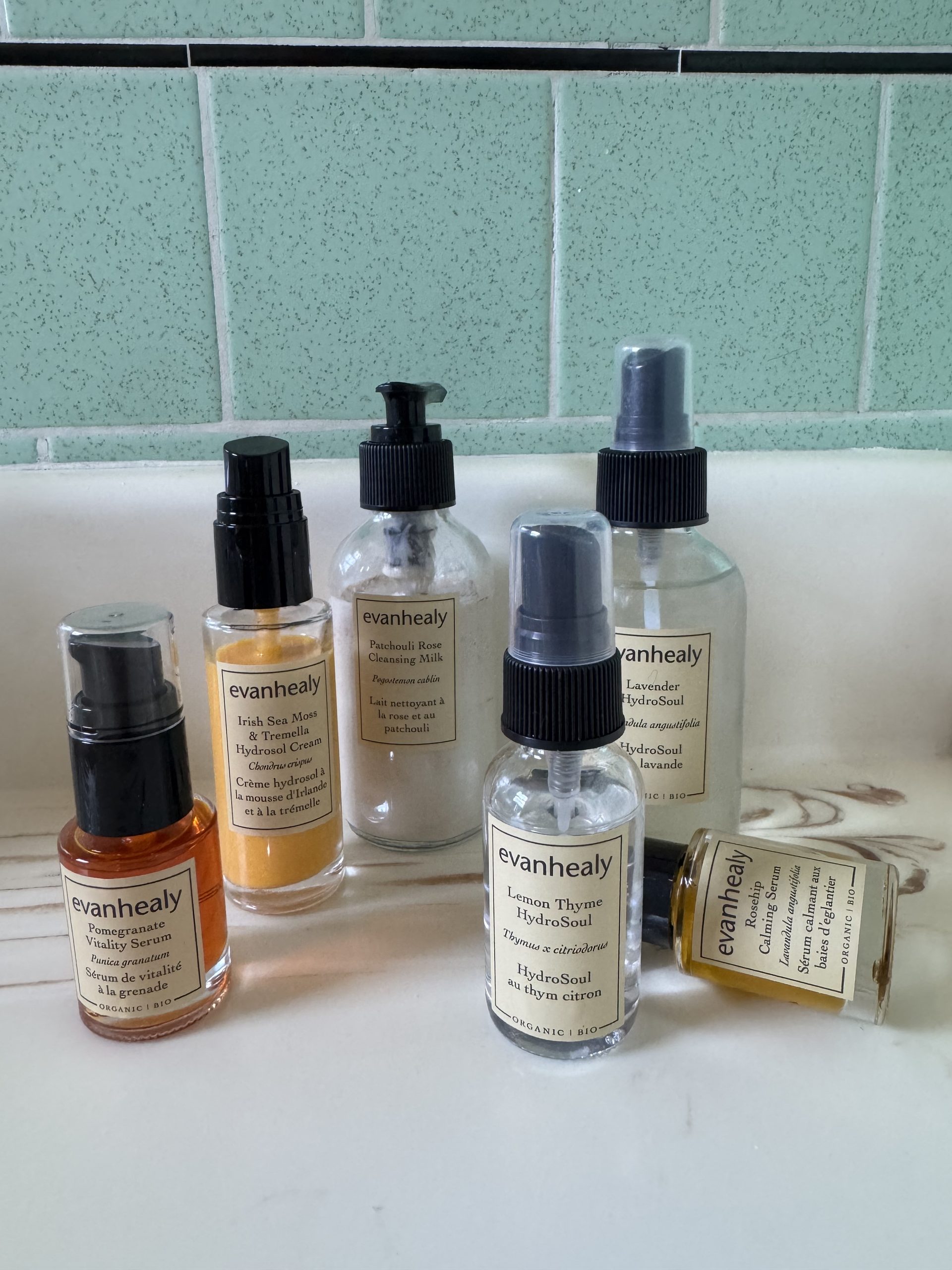 Six Evanhealy skincare bottles, including serums, cleansing milk, and hydrosols, arranged on a bathroom counter.