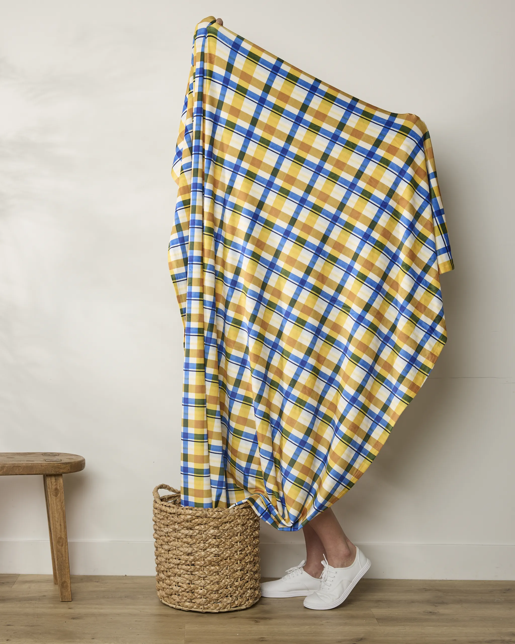 A person standing behind a large piece of yellow, blue, and white plaid fabric, wearing white sneakers.