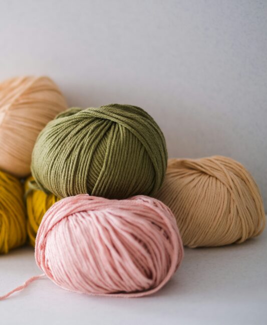 Four balls of yarn in pink, green, beige, and yellow are stacked on a white surface with a light background.