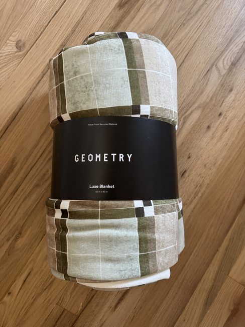 A rolled, plaid-patterned Geometry Luxe Blanket with a product band reading “Made From Recycled Material,” “GEOMETRY,” and “60 in x 80 in.”