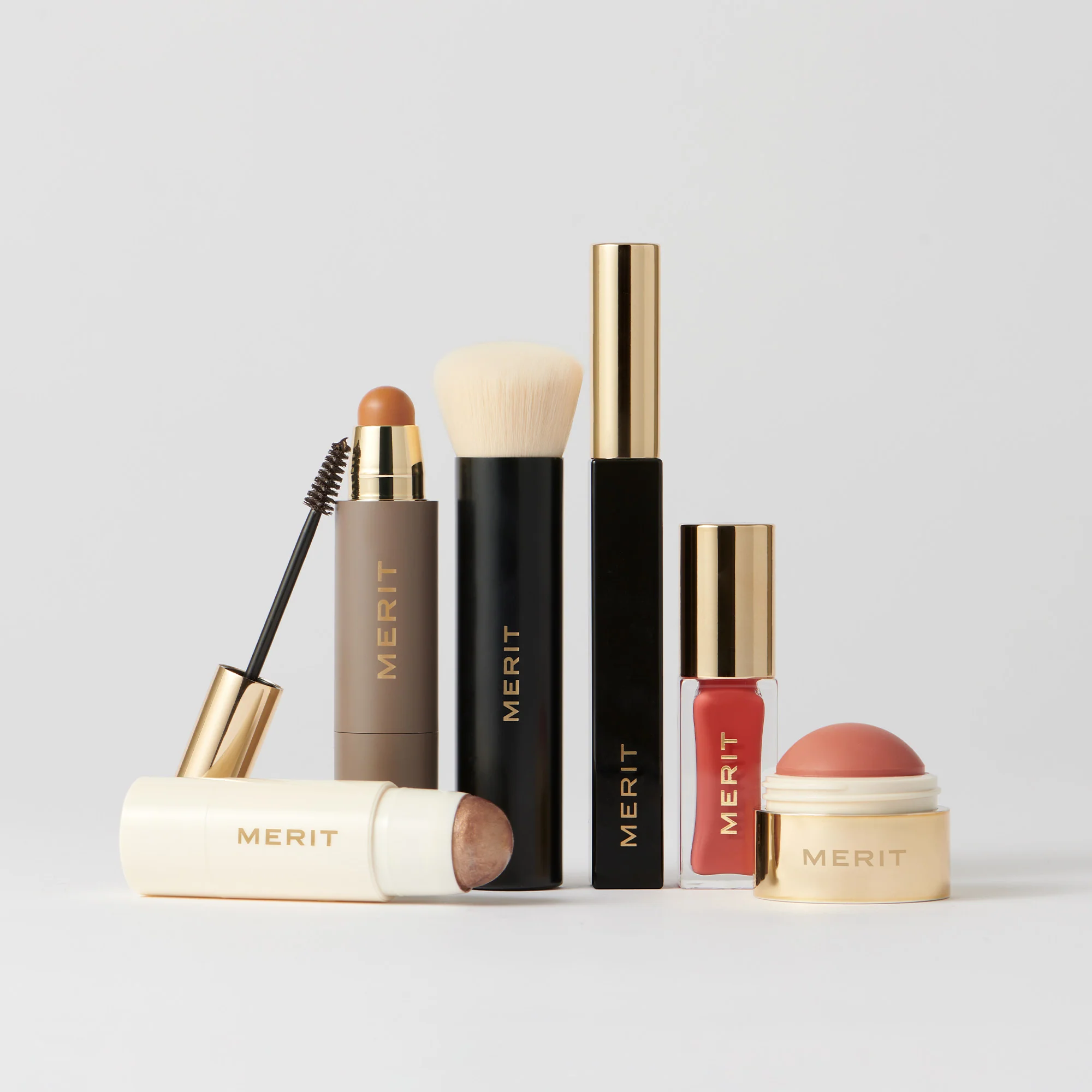 A collection of Merit makeup products, including brushes, lip color, mascara, and highlighter sticks, arranged against a white background.