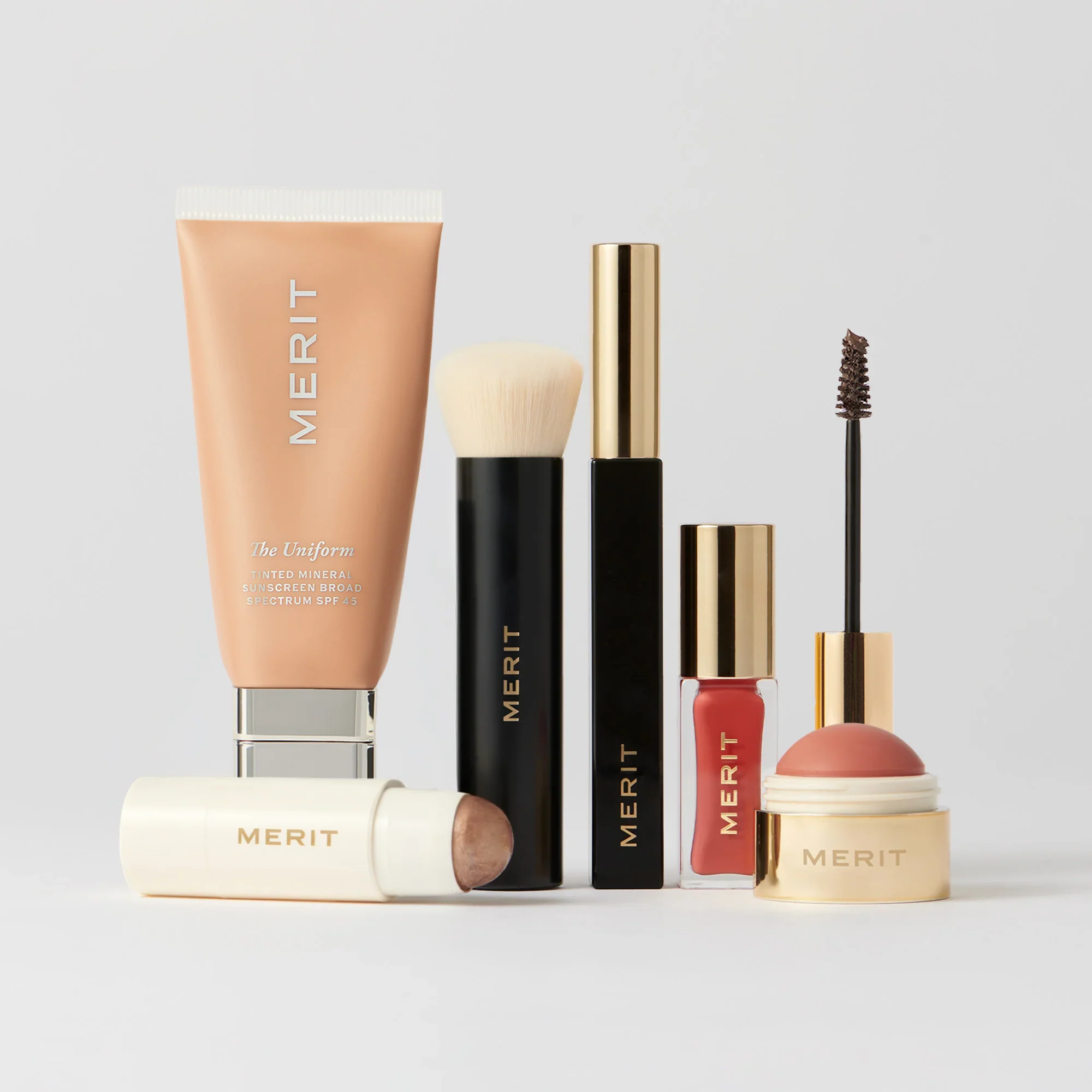 A collection of Merit makeup products, including a tube of tinted sunscreen, a highlighter stick, a makeup brush, mascara, lip oil, and a blush balm.
