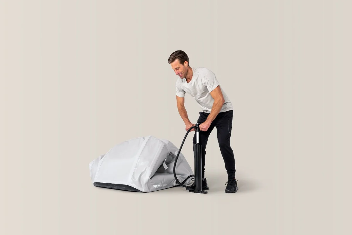 A man uses a hand pump to inflate a gray, folded tent on a light background.