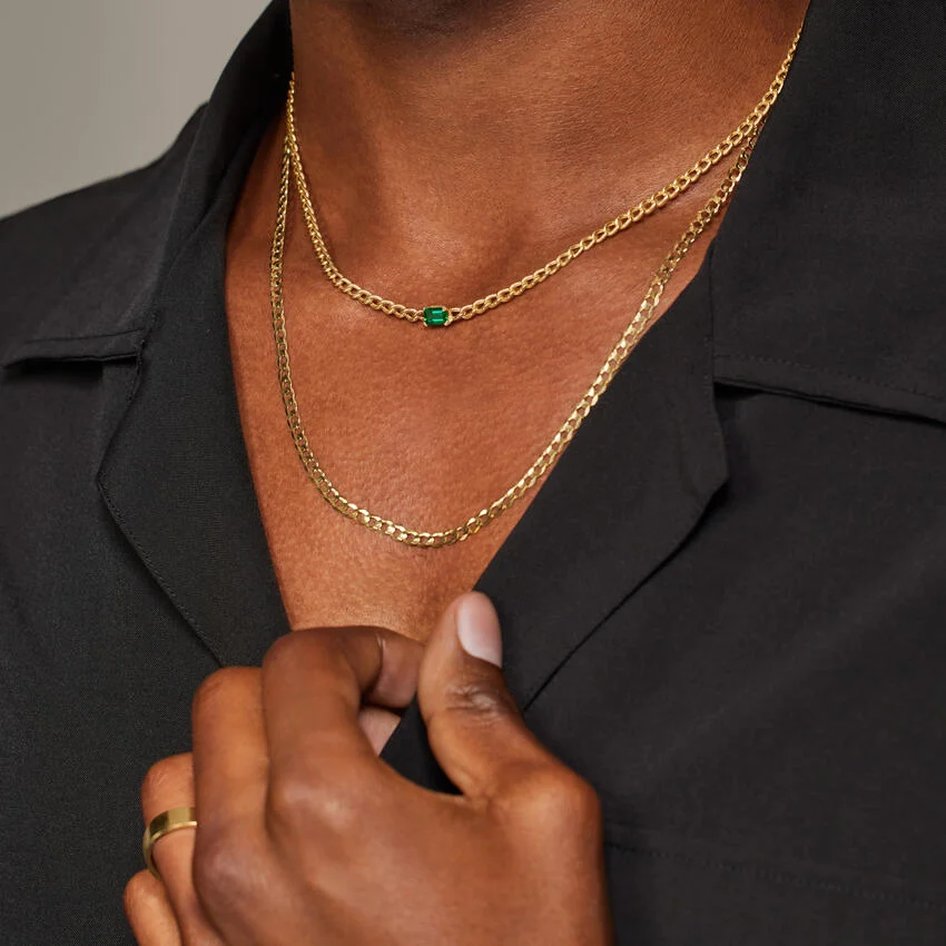 A person wearing two gold chain necklaces, one featuring a small emerald pendant.