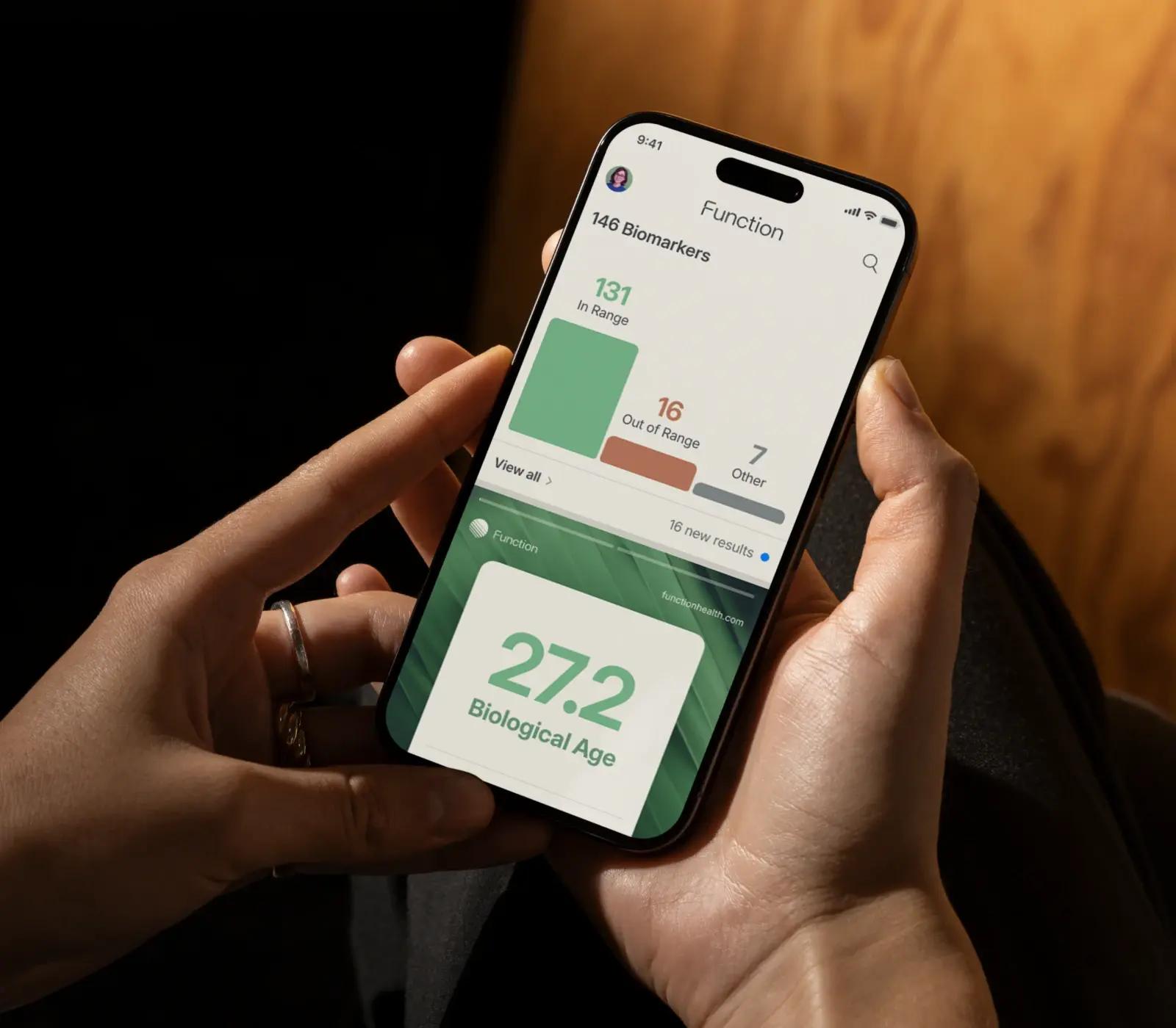 A person holds a smartphone displaying the Function health app, which shows biomarker statistics and a biological age of 27.2.