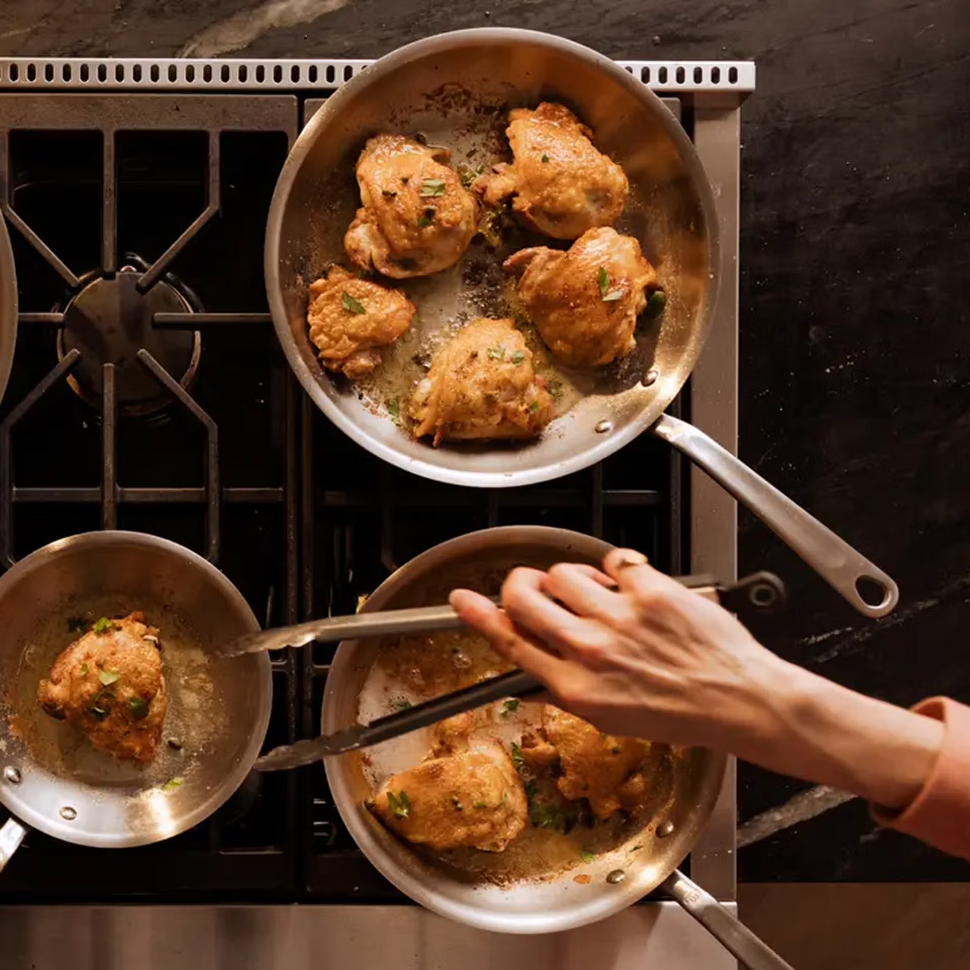 A person uses tongs to turn chicken thighs searing in three stainless steel pans on a gas stovetop.