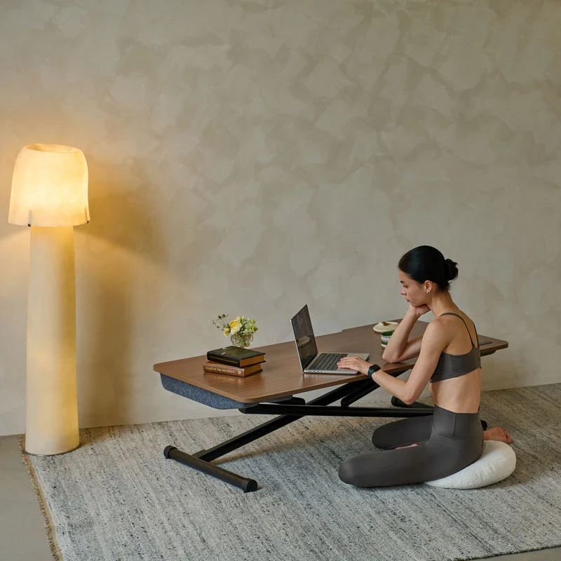 A person sits cross-legged on a floor cushion, working on a laptop at a low-profile adjustable desk next to a floor lamp.