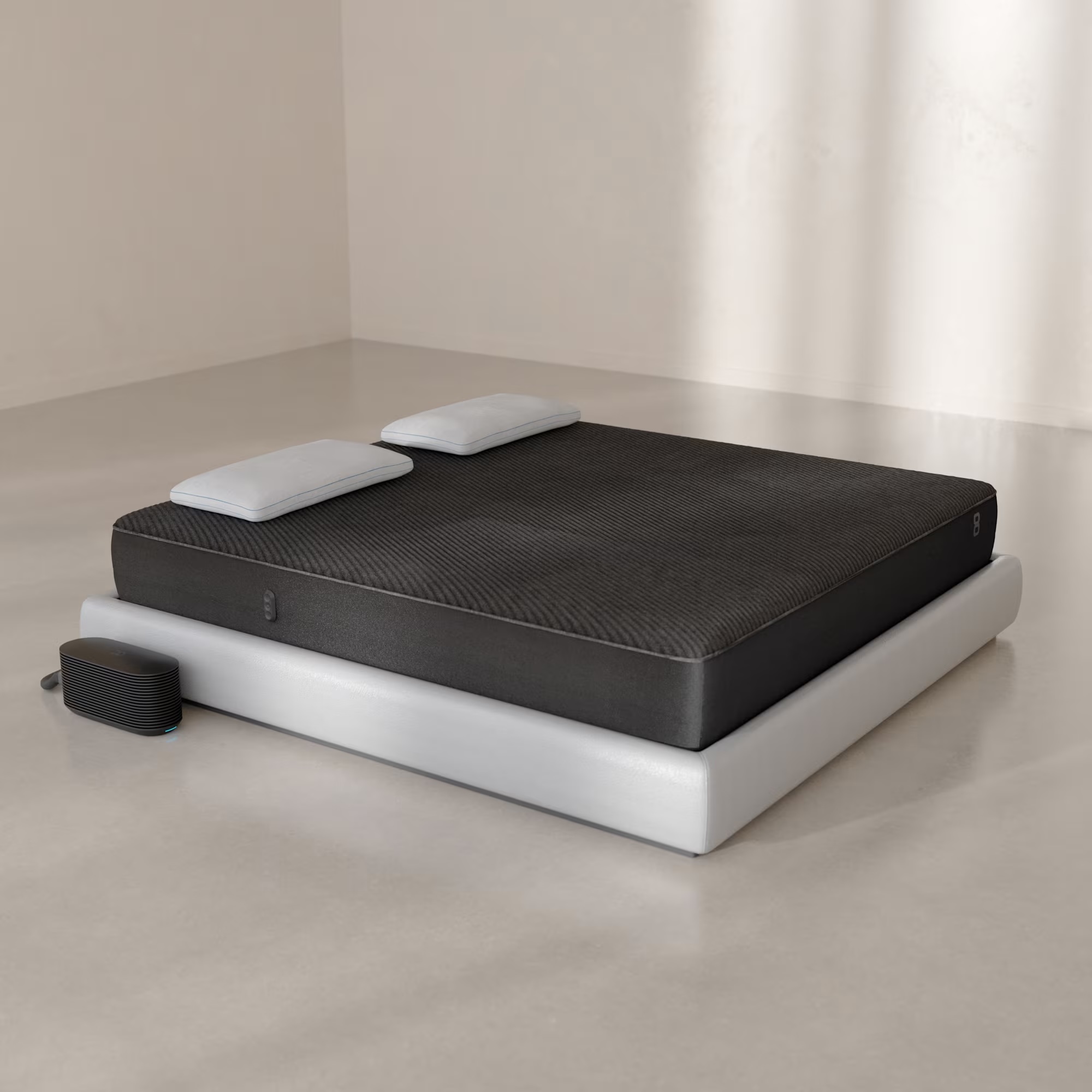 A dark mattress on a white platform bed frame with two pillows and a small bedside device on the floor.