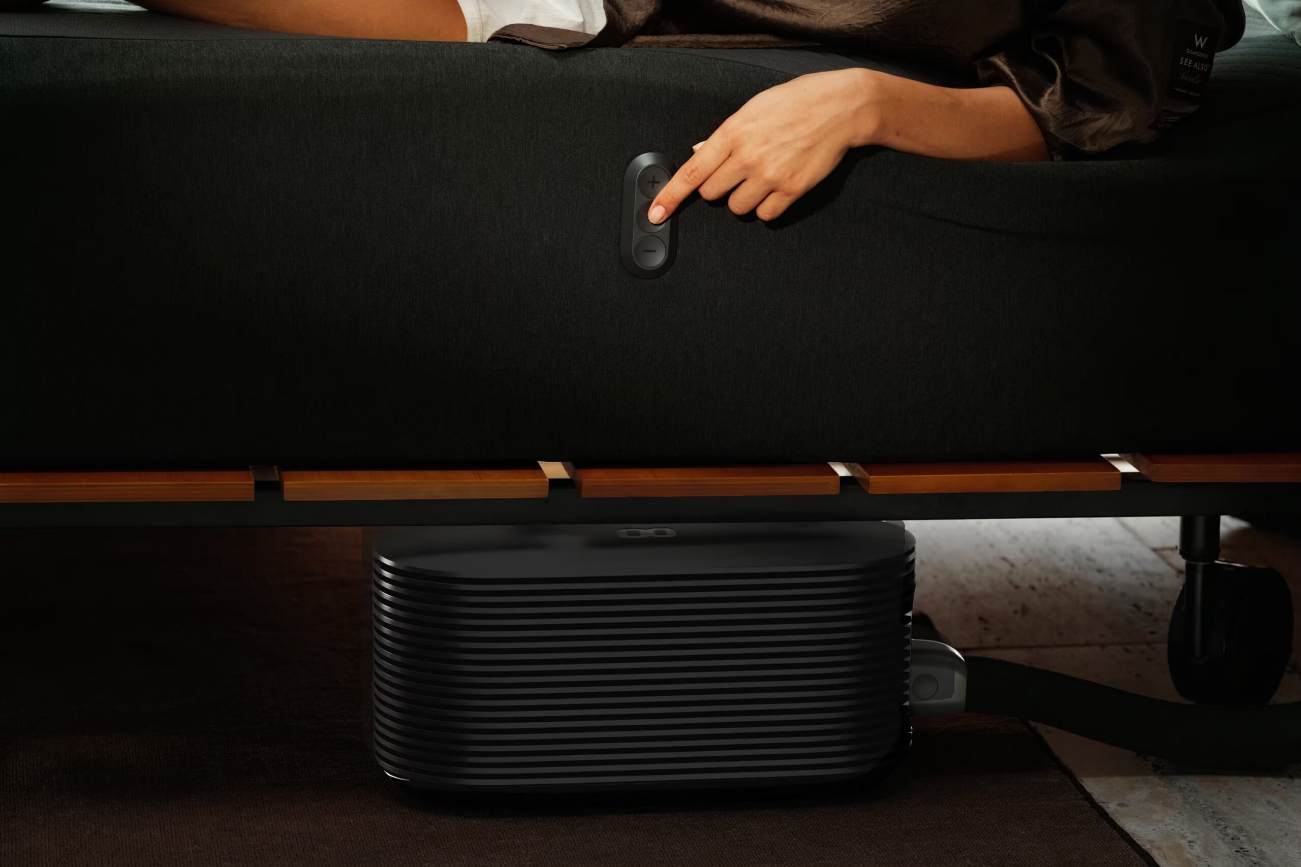 A person’s hand presses a button on the side of a bed frame, which is connected to a black rectangular air climate control unit positioned underneath.