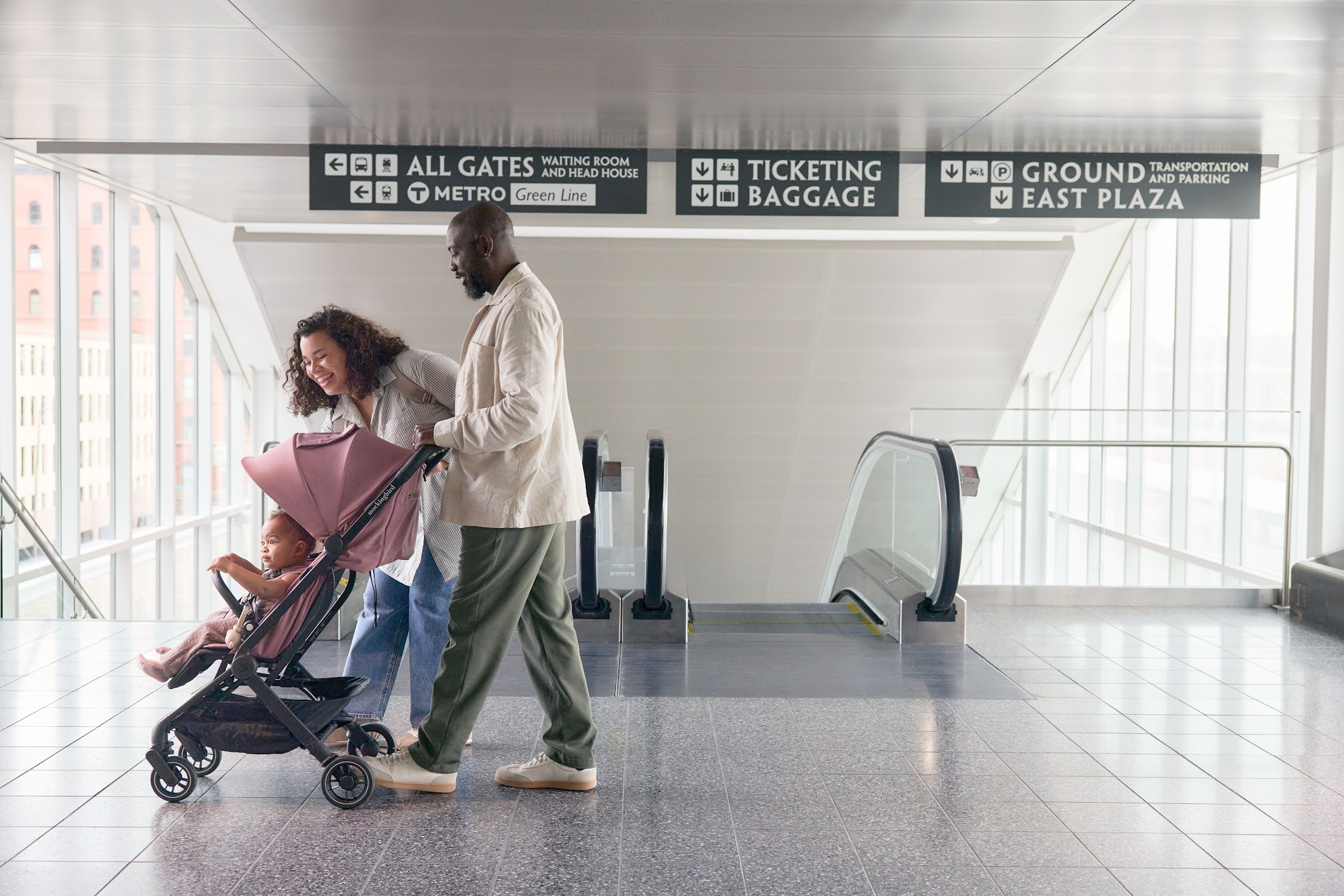 A man and a woman push a toddler in a stroller through a train station concourse with overhead signs for gates, ticketing, and ground transportation.