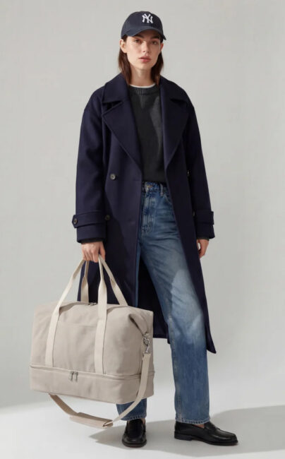 A woman wearing a dark blue coat, jeans, and a baseball cap holds a large beige canvas duffel bag.