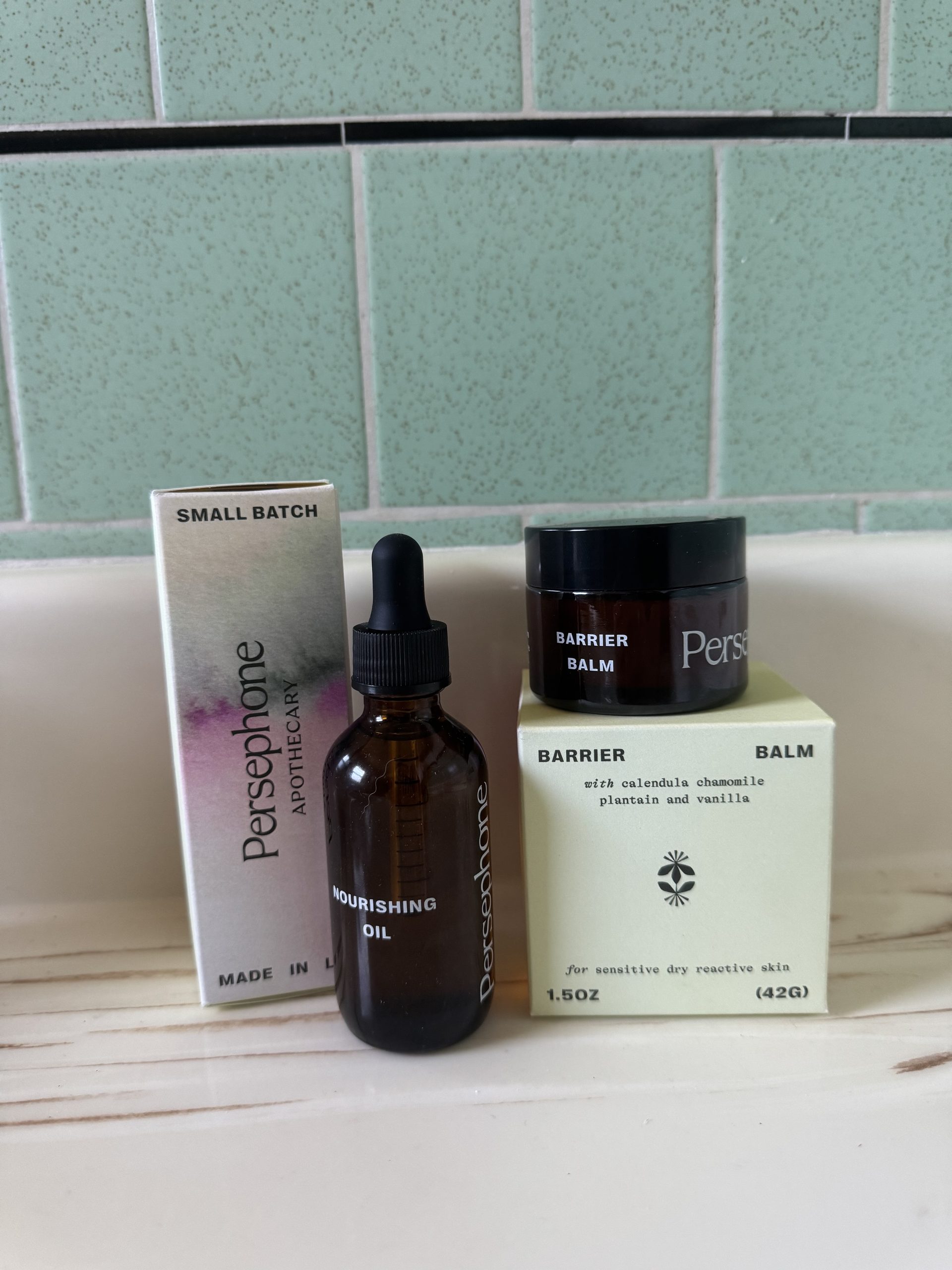 Persephone Apothecary Nourishing Oil in an amber dropper bottle with its box, and Barrier Balm in a jar with its box, sitting on a counter.