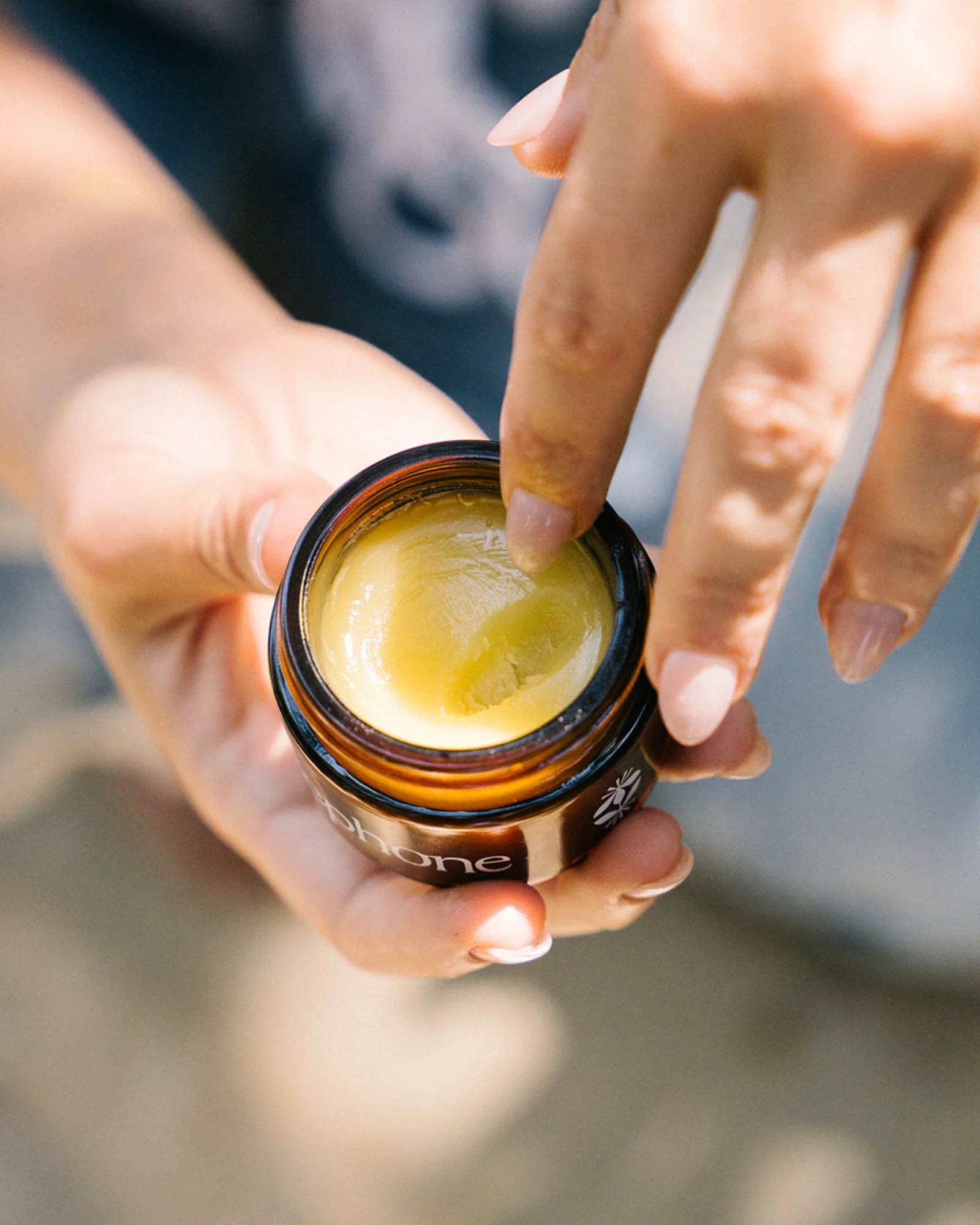A finger scoops a small amount of yellow balm from a glass jar.