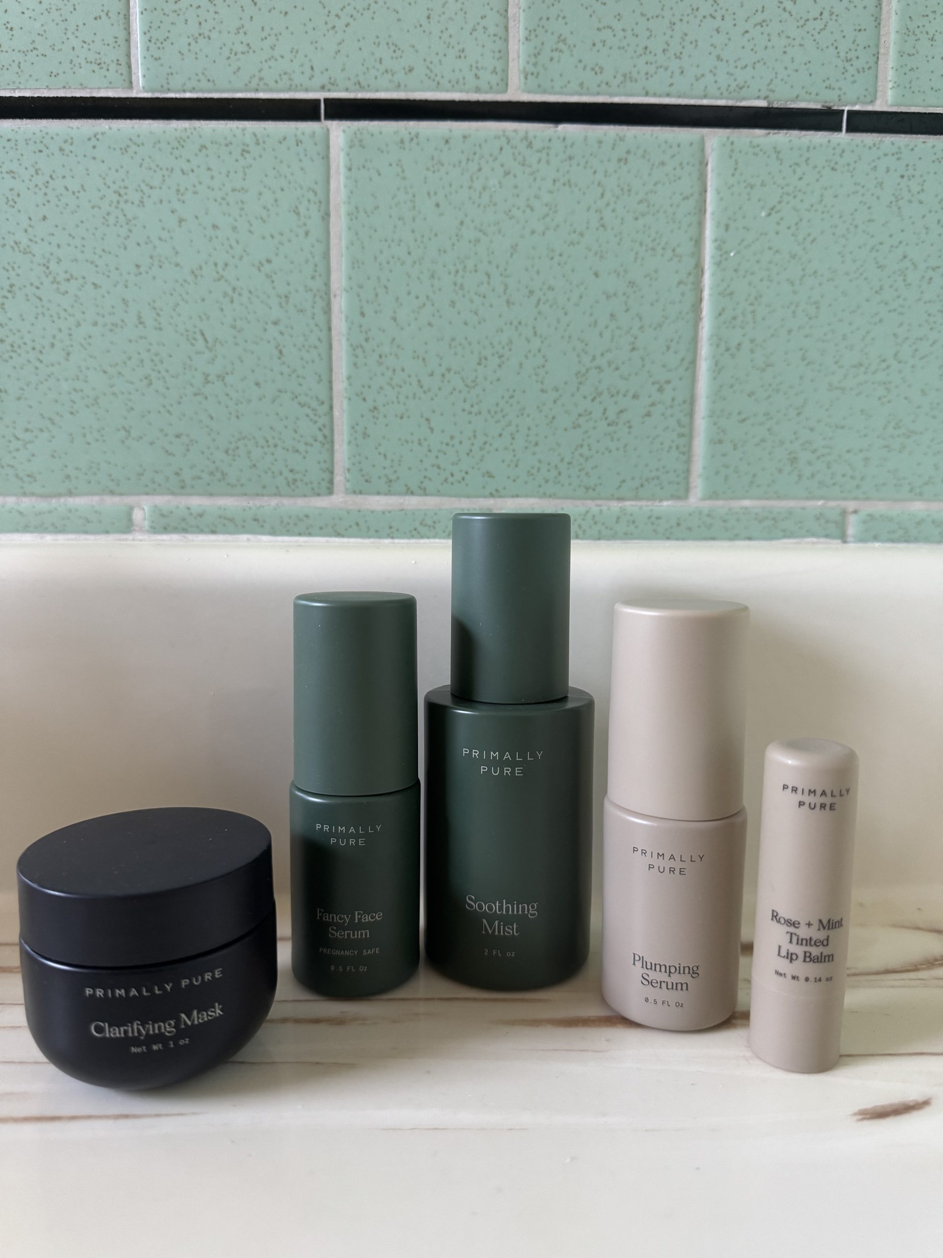 Five Primally Pure skincare products including a jar of Clarifying Mask, Fancy Face Serum, Soothing Mist, Plumping Serum, and Rose + Mint Tinted Lip Balm.