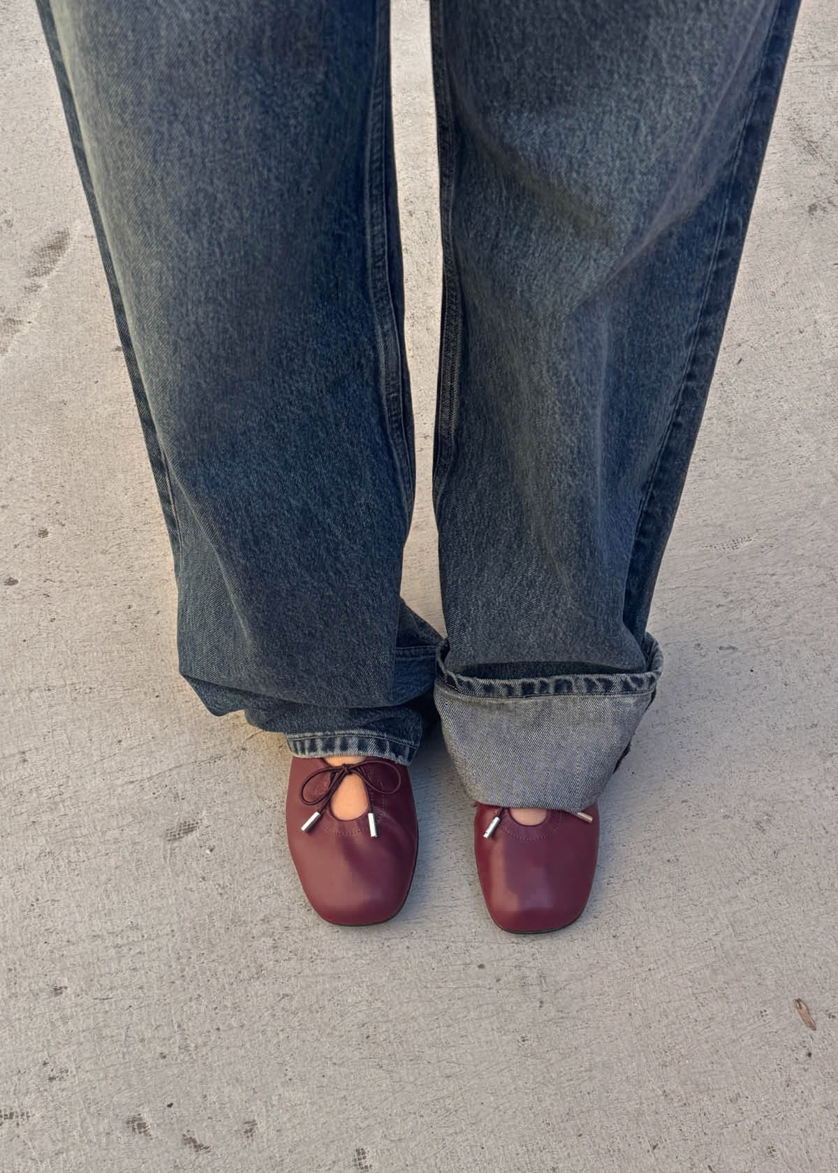 A person wearing loose-fit blue jeans and maroon ballet flats with tied cord details.