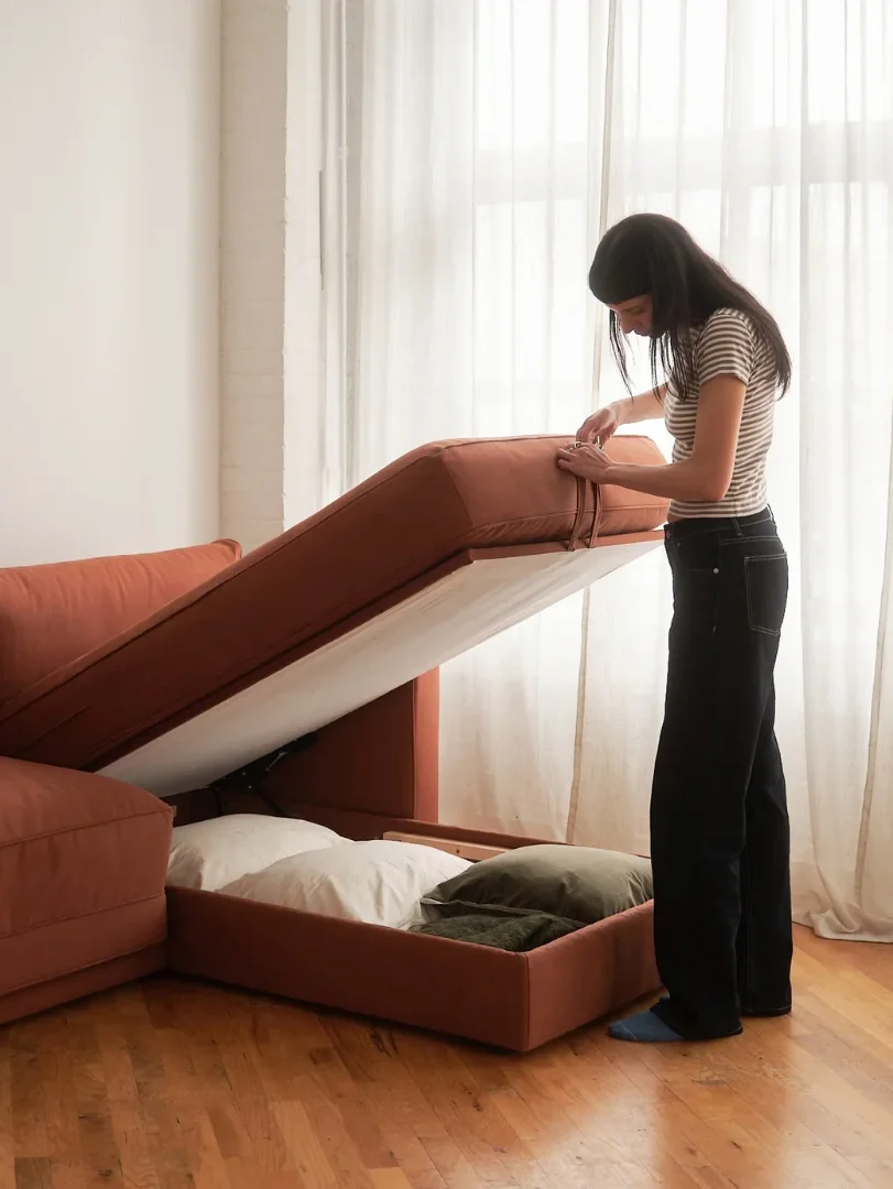 A person lifts the seat of a rust-colored sectional sofa to reveal hidden storage space underneath.
