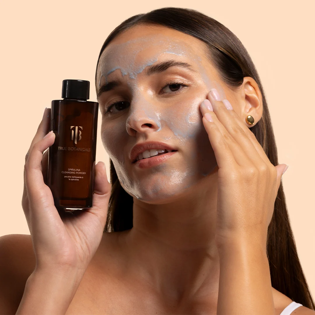 A person applying blue cleansing powder to their face while holding a bottle of True Botanicals Spirulina Cleansing Powder.
