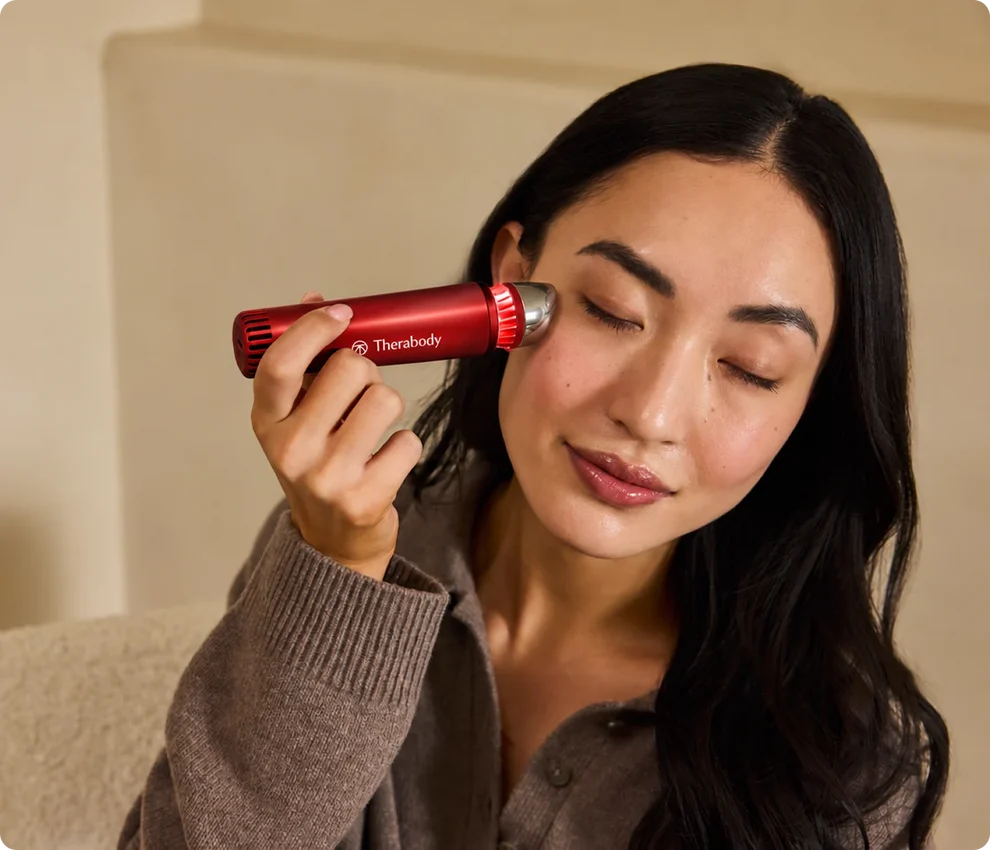 A person uses a red Therabody handheld facial device on their cheek.
